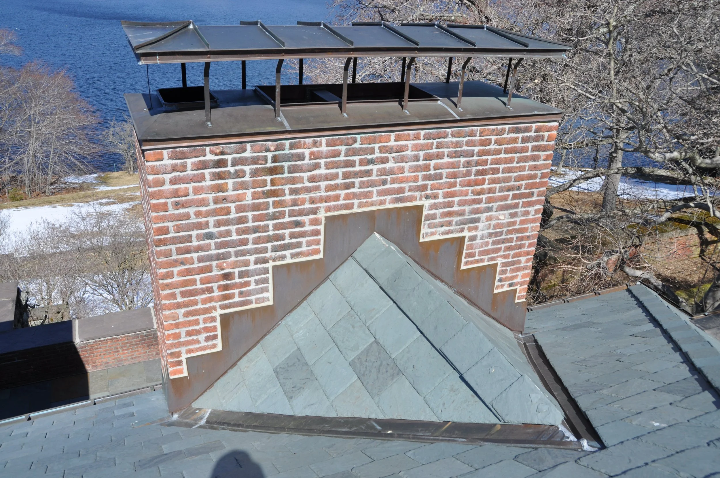 A brick chimney with a metal cap and a sloped metal roof below it, seen from above. There are leafless trees and a body of water in the background, indicating a winter setting.