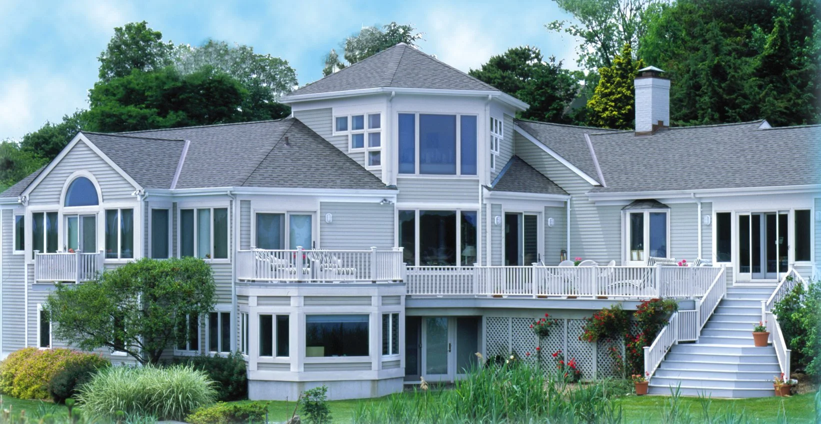 A large modern house with multiple levels, gray siding, and a multi-pane bay window. It features a spacious deck with white railings, outdoor furniture, and stairs leading down to a landscaped yard with greenery and flowering plants. The house is surrounded by tall trees under a partly cloudy sky.