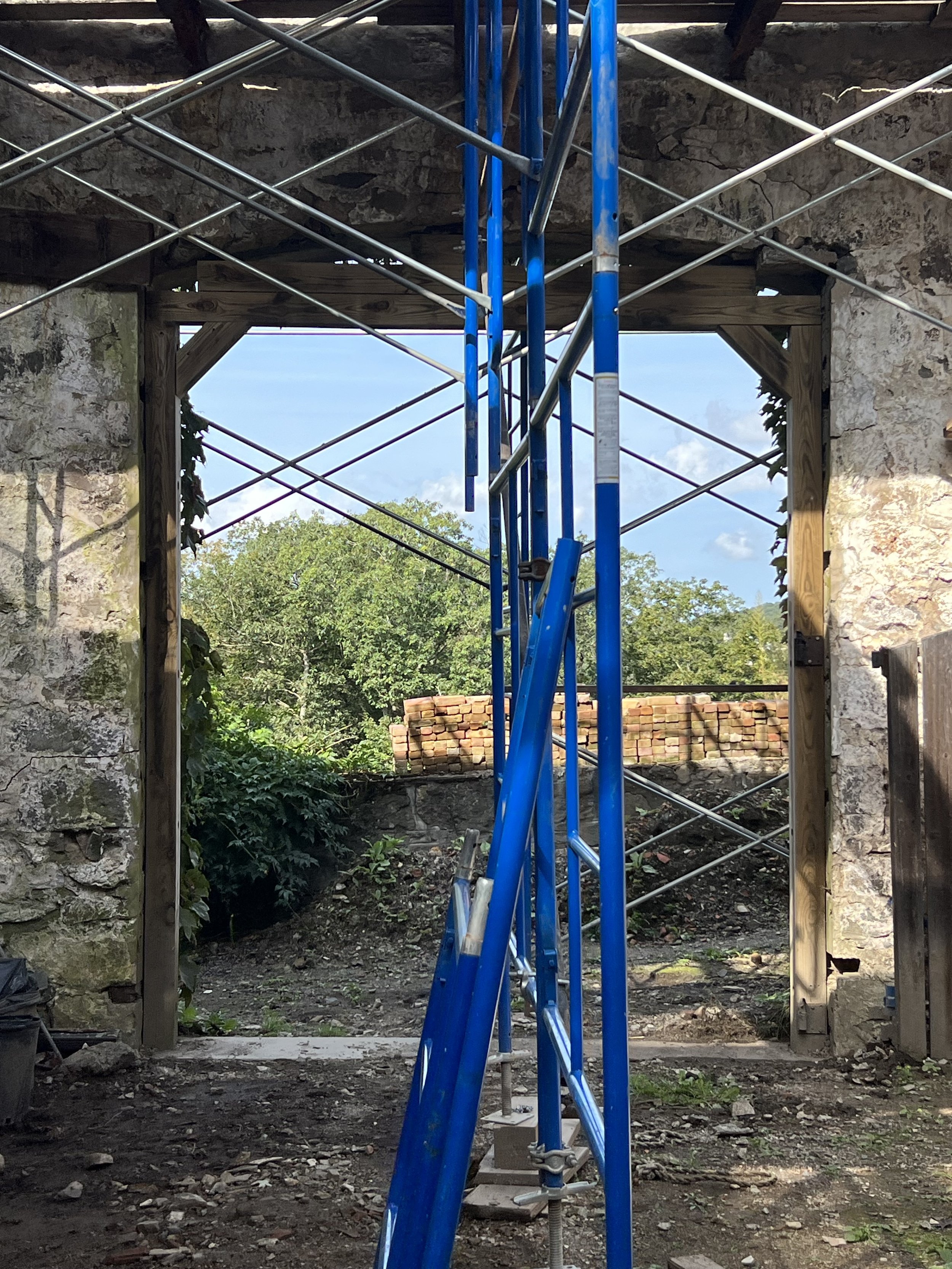 A construction site with blue scaffolding set up inside a building with stone walls. The area is framed by wooden beams, and outside the space, there is greenery and a stack of bricks visible in the distance.
