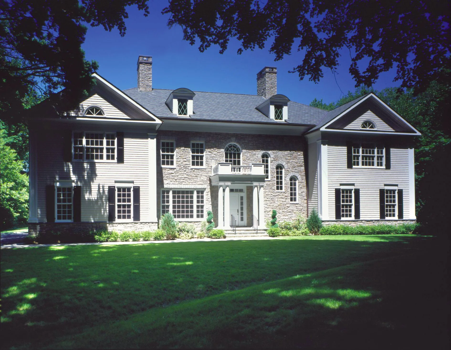 A large two-story house with stone and beige siding exterior, multiple windows with black shutters, a covered front entrance with steps, surrounded by a manicured lawn, trees, and shadows cast on the grass, under a clear blue sky.