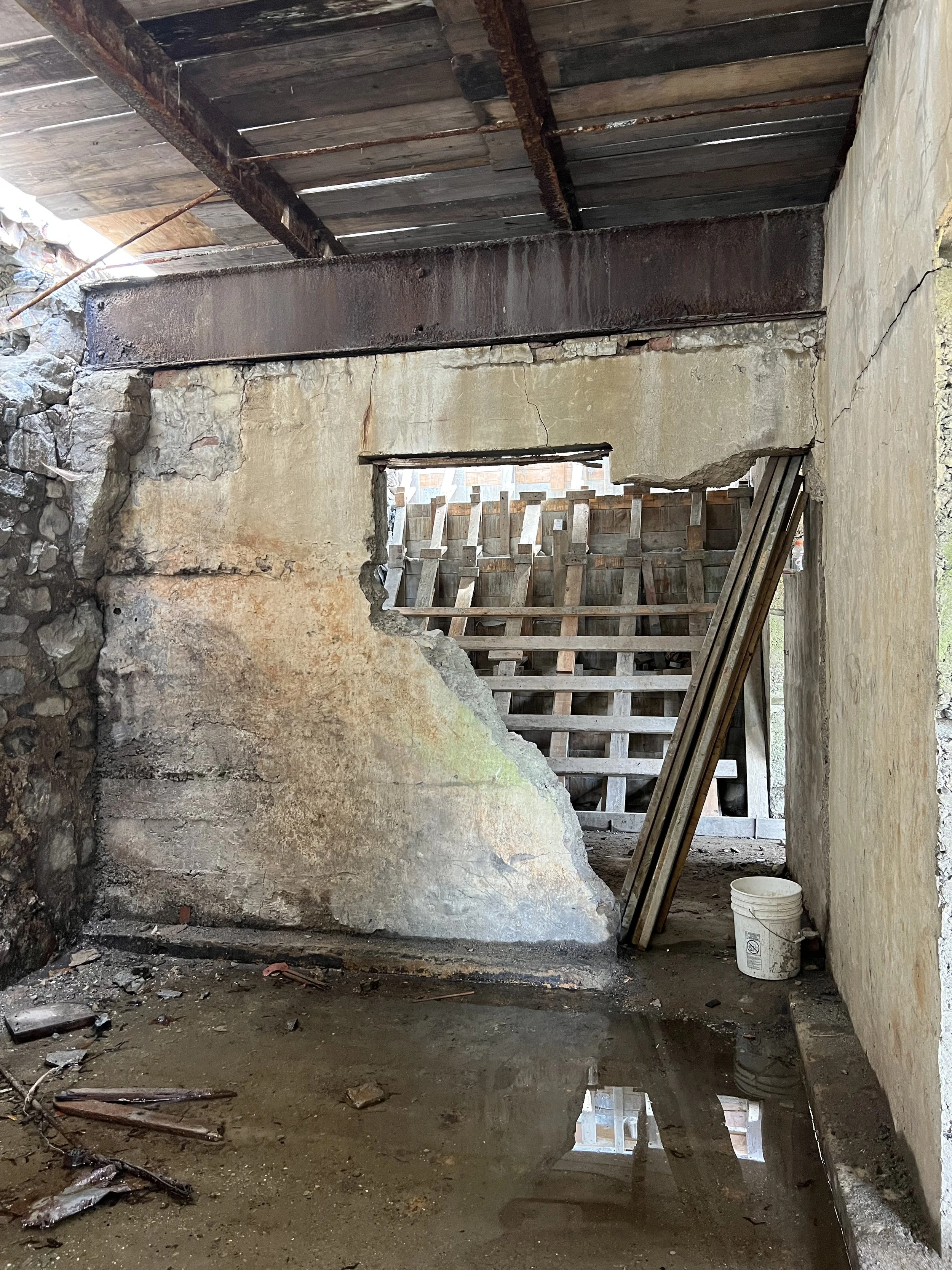 A construction site with a partially demolished concrete wall, wooden beams on the ceiling, and a muddy floor with debris. There is a white bucket and some wooden planks leaning against the wall, with sunlight coming through gaps in the roof and a small window.