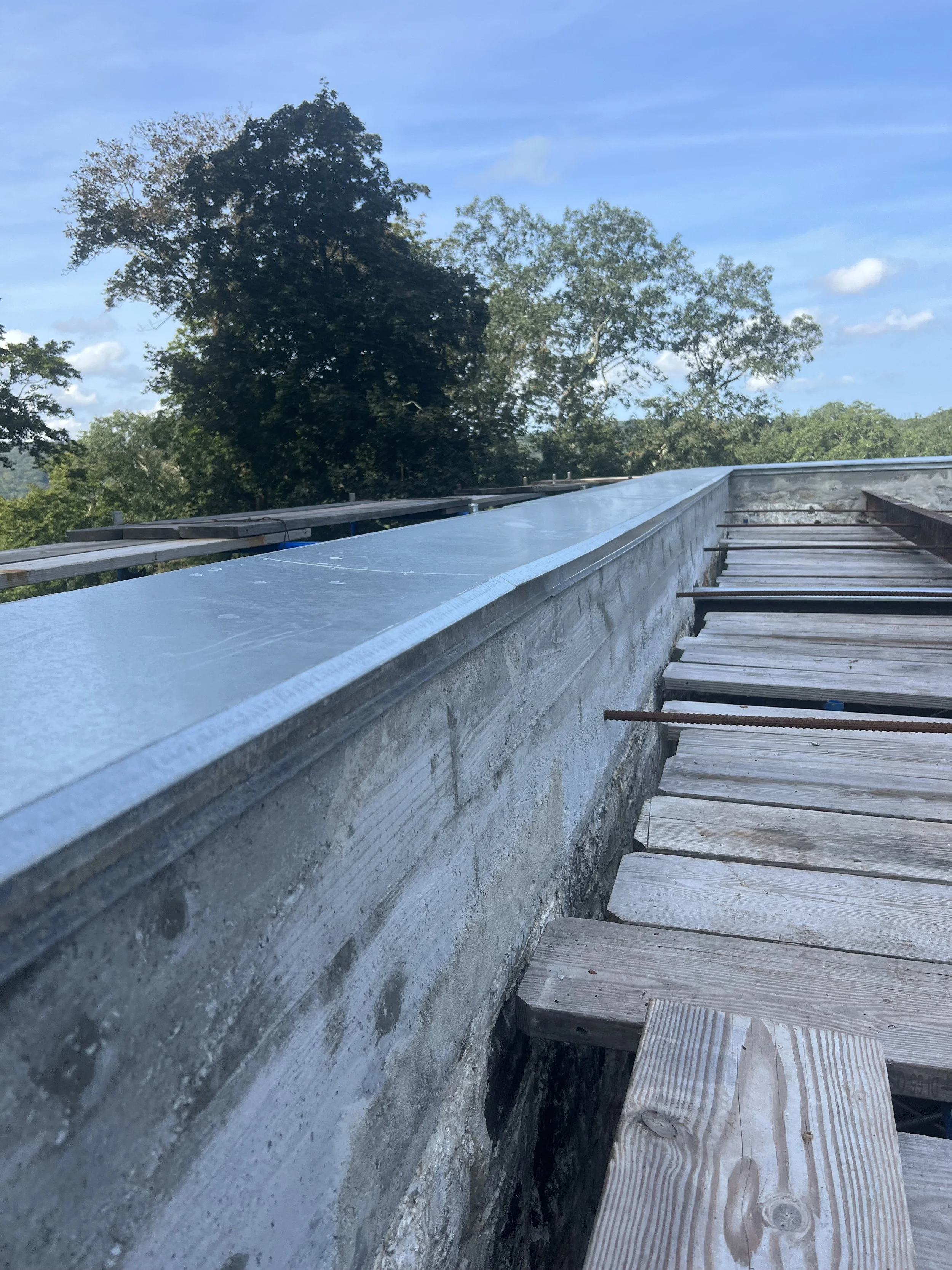 Construction site with a concrete wall and wooden planks, trees, and blue sky in the background.