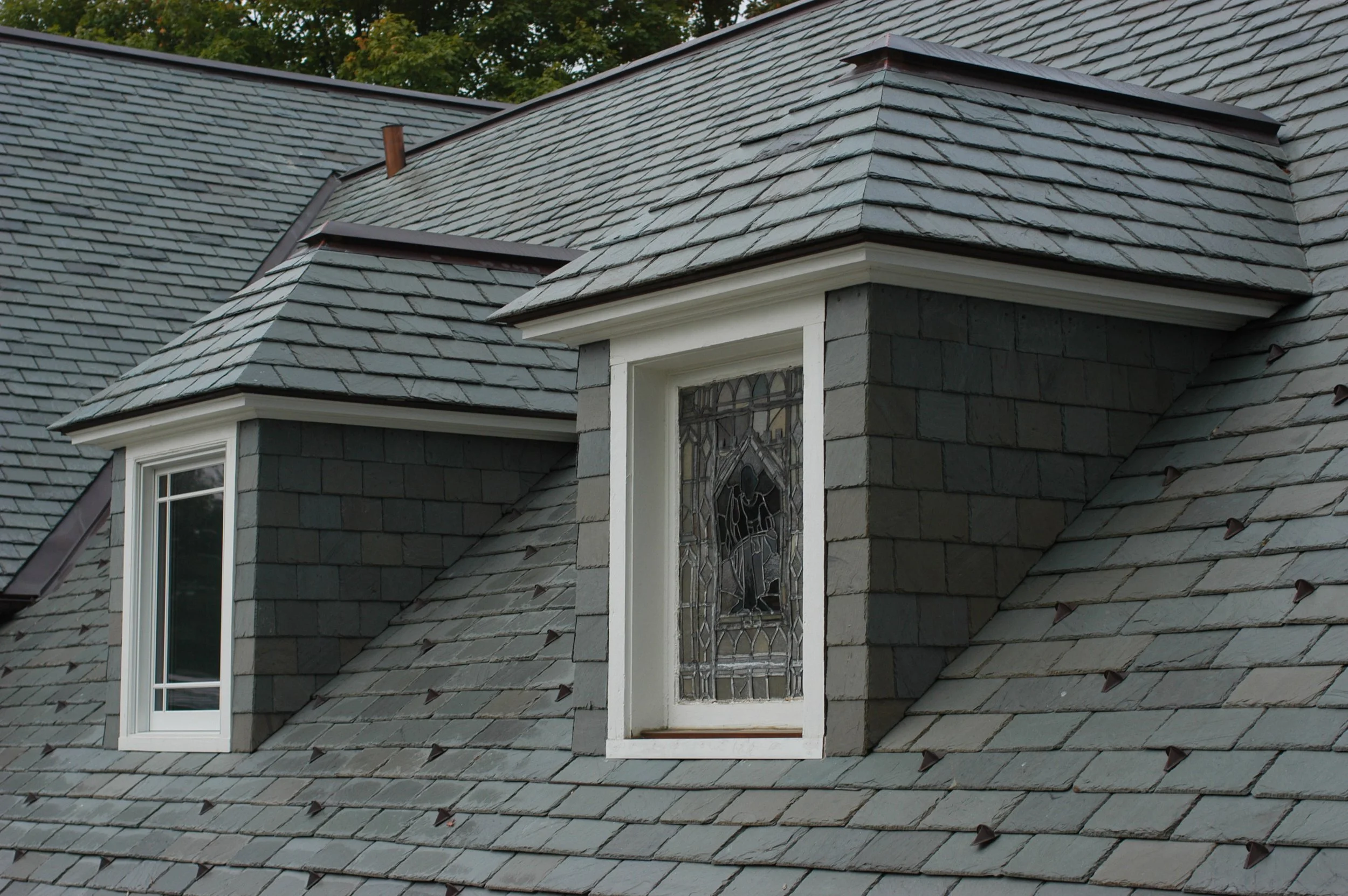 Close-up of a house roof with slate shingles, featuring a skylight window with stained glass and a section of a dormer window.