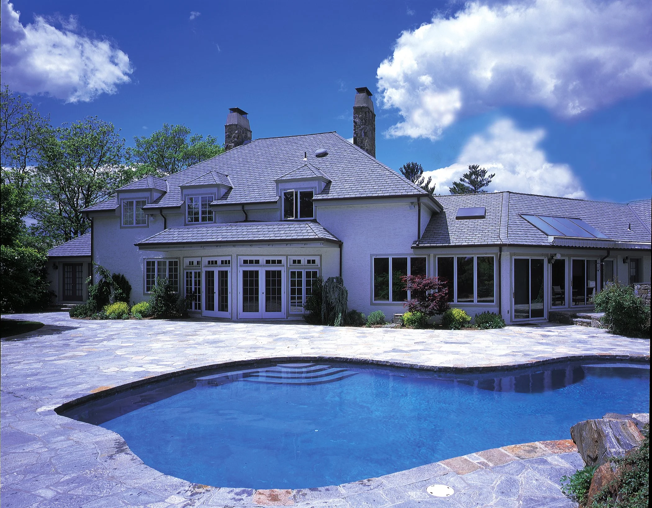A large modern house with a swimming pool in the backyard. The house has multiple chimney stacks, large windows, and a sloped roof. The backyard is landscaped with plants and trees, and the pool is surrounded by stone paving.