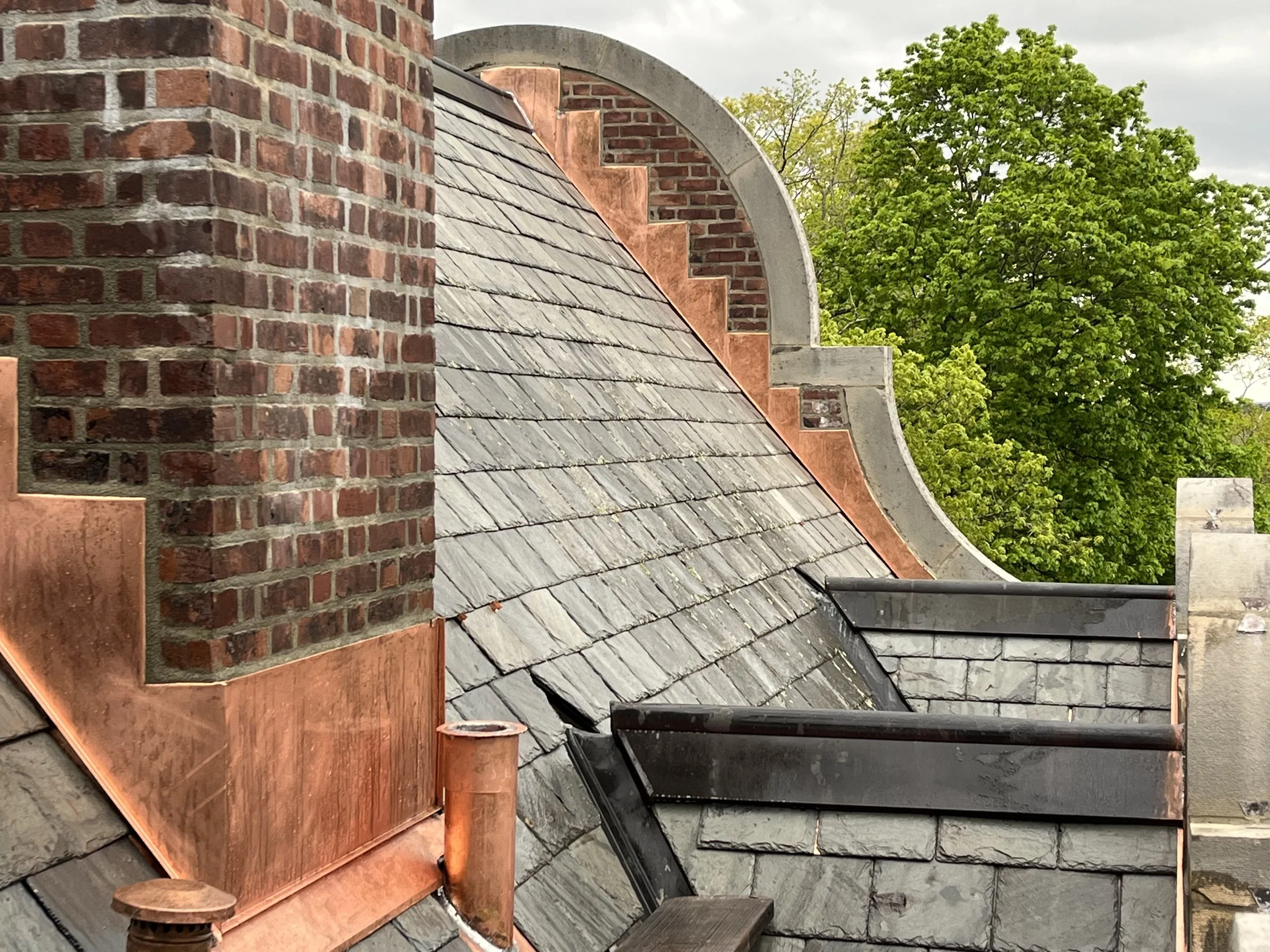 Roof with slate shingles, brick chimney, and trees in the background.
