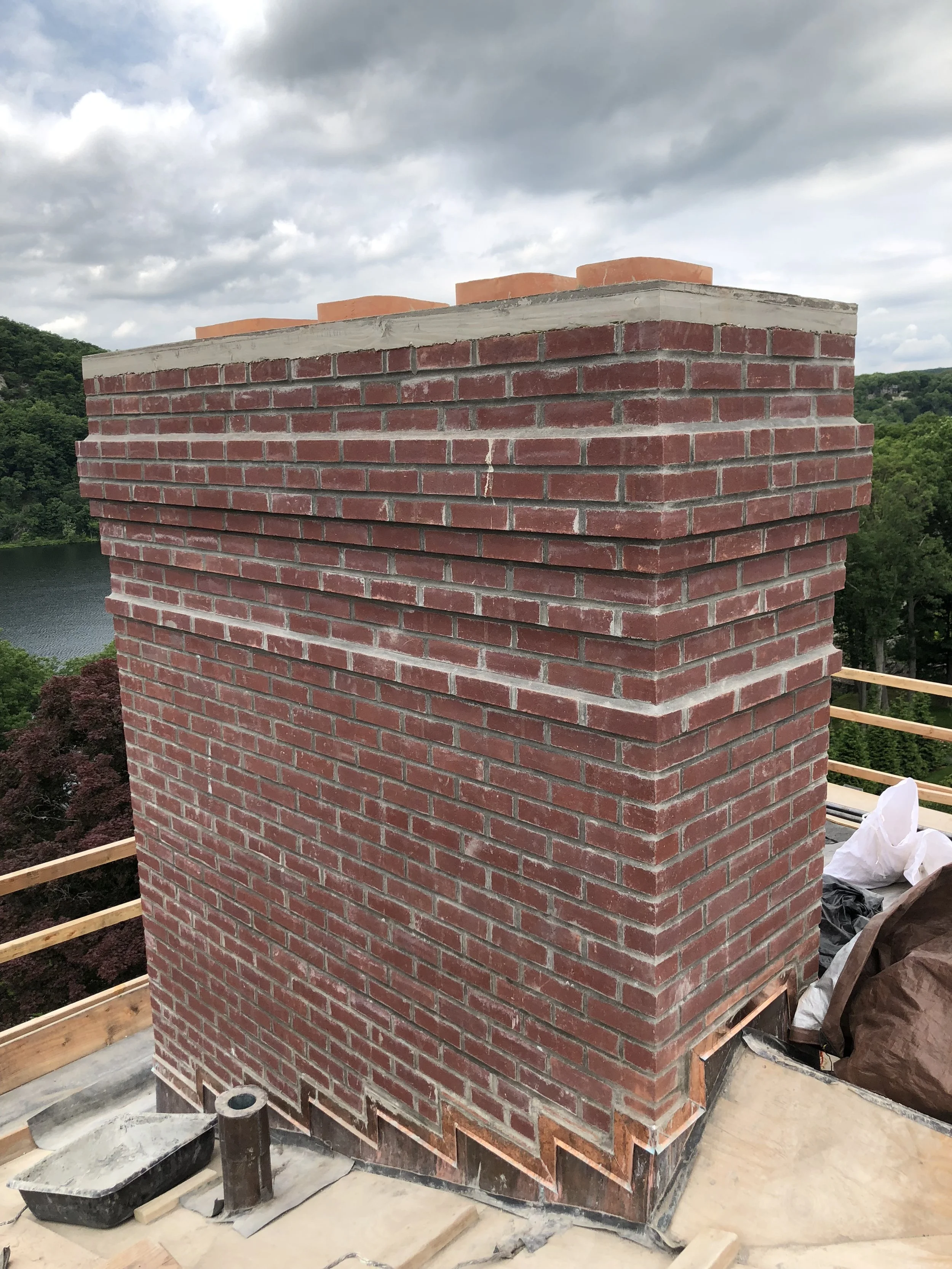 A brick chimney under construction on a building, with a scenic view of trees and a lake in the background, and cloudy sky overhead.