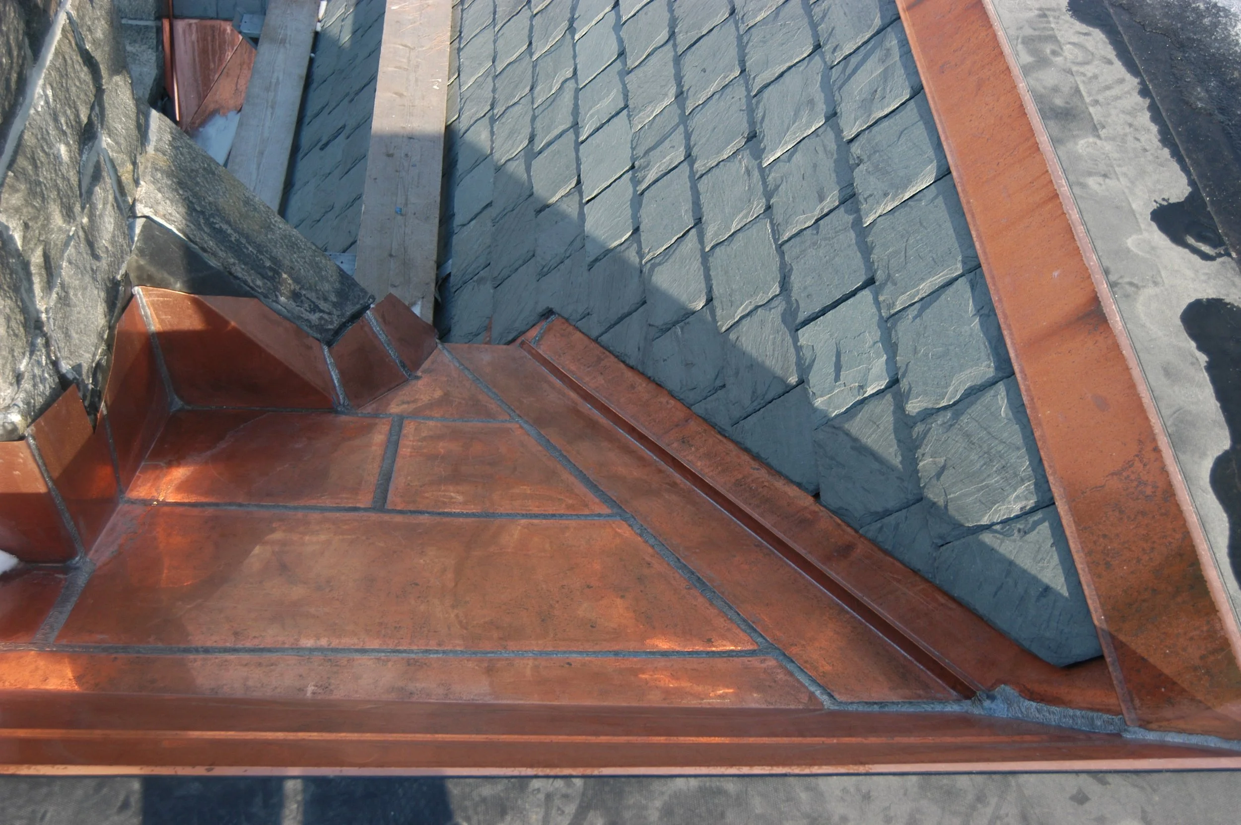Close-up of a slate roof with red clay tiles along the edges, showing the textured gray slate shingles and part of a chimney made of similar slate material.