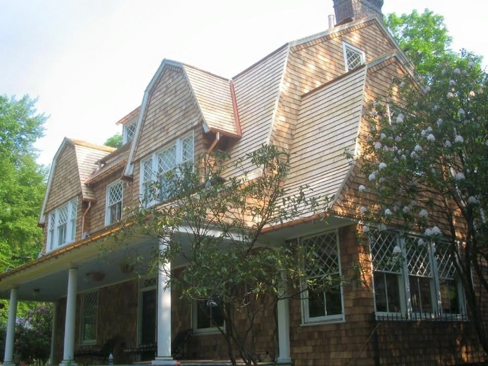 A large Victorian-style house with a complex roof design, wooden shingles, multiple windows, and a front porch with white columns, surrounded by trees.