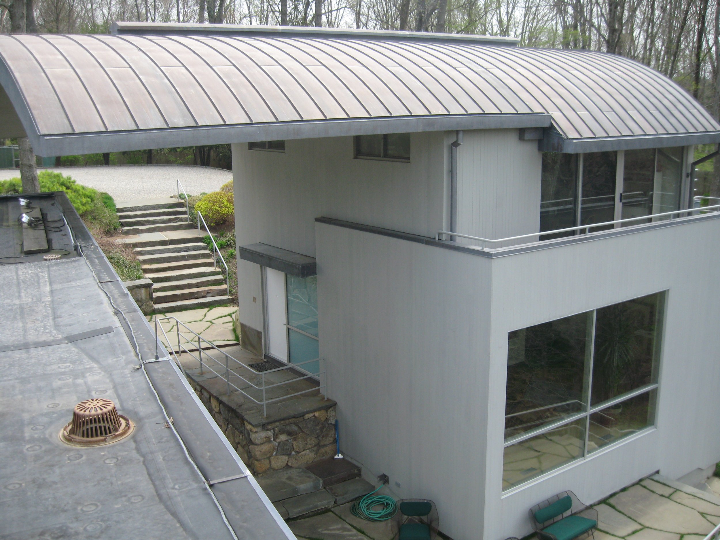 A modern house with a curved metal roof, large windows, and a small staircase leading to a patio. The house is surrounded by trees and greenery.