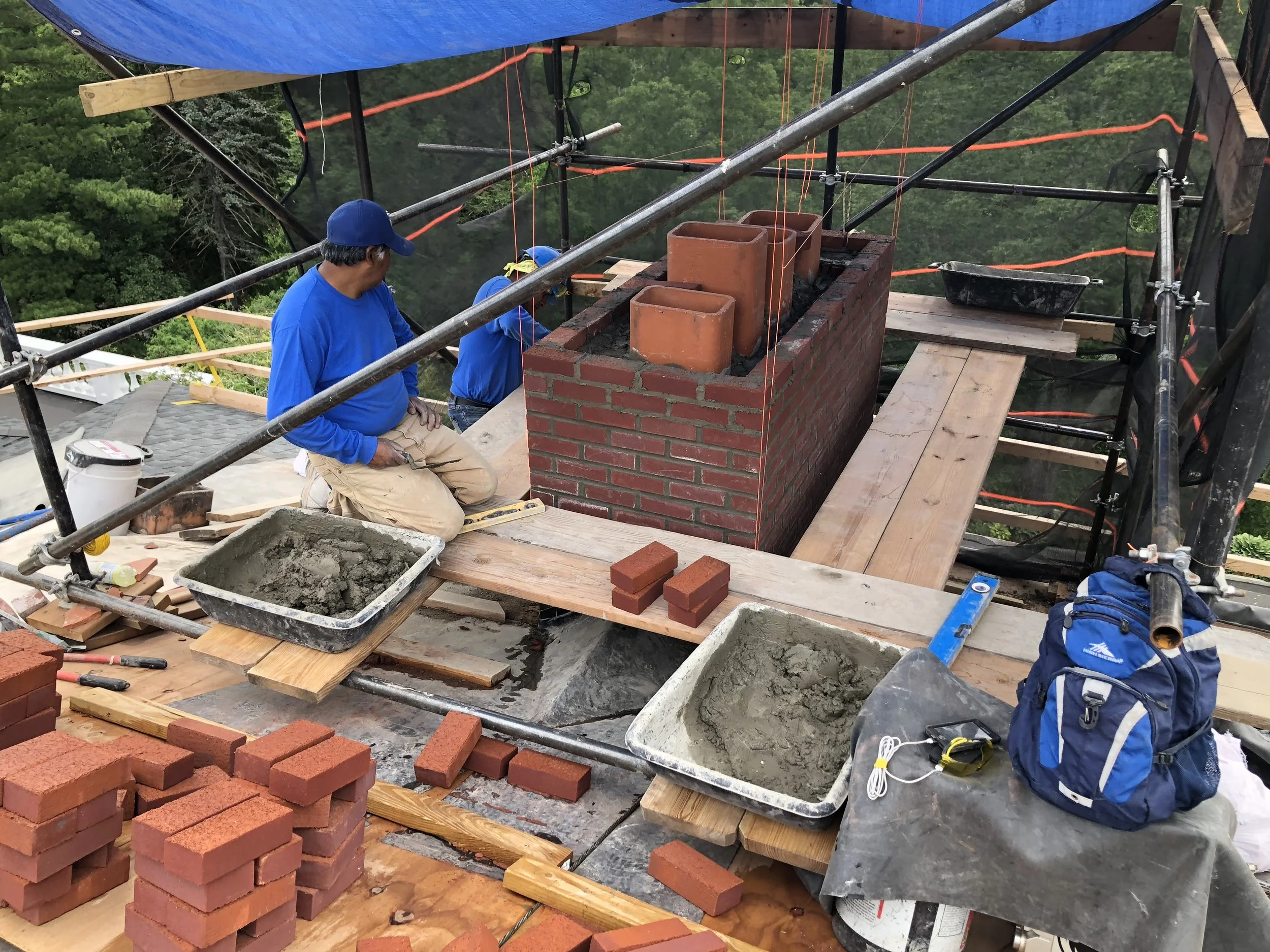 Construction workers building a brick chimney on a rooftop, surrounded by scaffolding and construction materials.