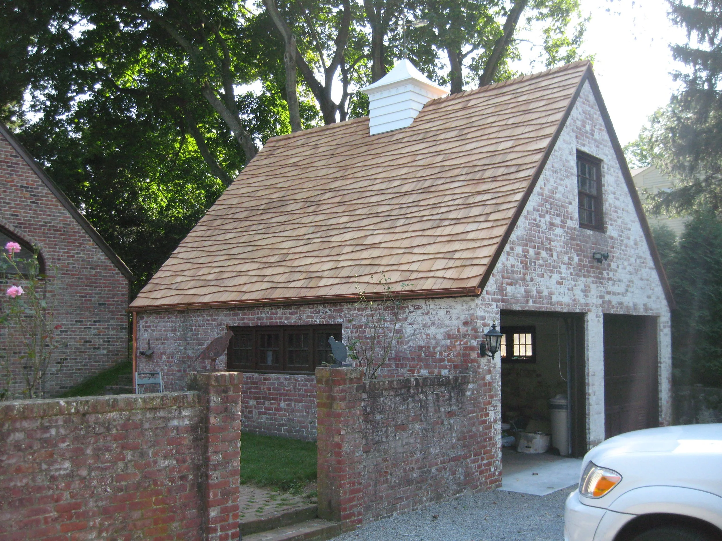 Brick house with a steep shingle roof, white chimney, and a window on the side. There is a brick wall with a small garden in front, and some trees in the background providing shade.