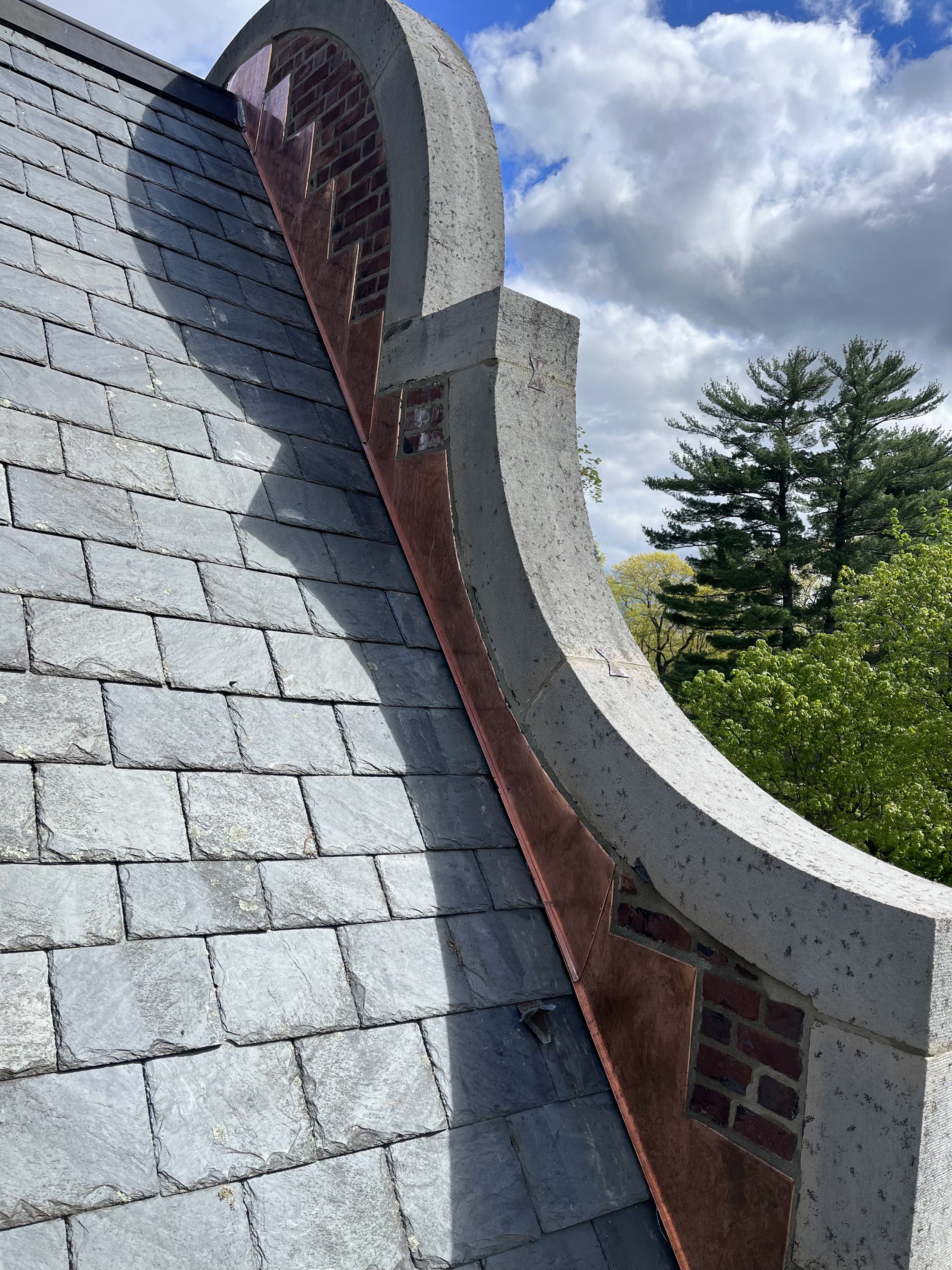 Close-up of a slate roof and decorative chimney with a copper trim, set against a backdrop of a cloudy sky and green trees.