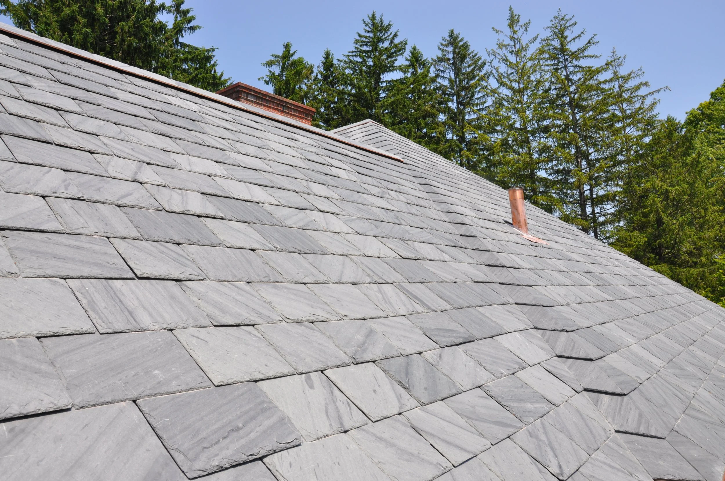 Slate roof with a chimney and surrounding trees under a clear blue sky.