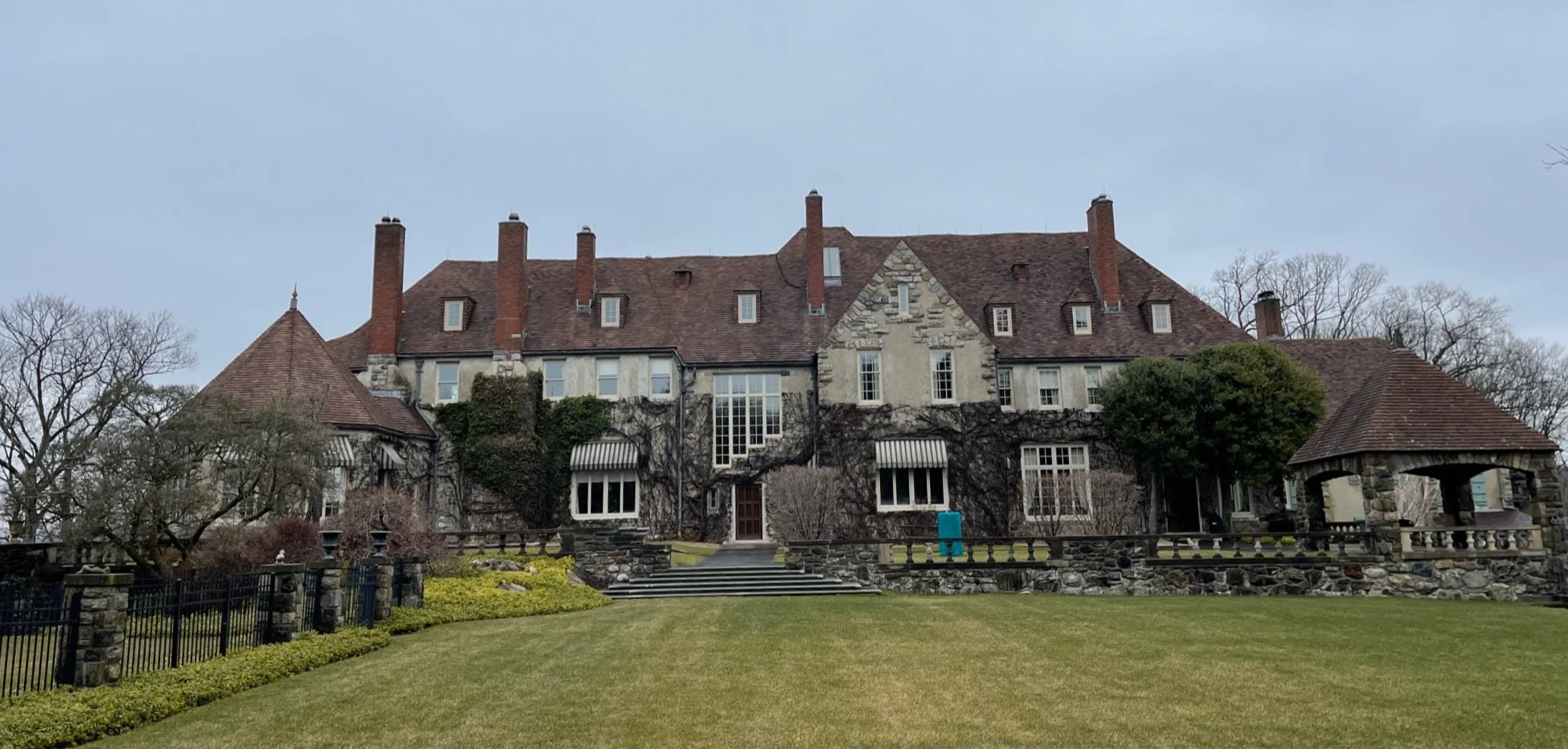 A large historic stone mansion with a steeply pitched roof, multiple chimneys, and ivy-covered walls, surrounded by a well-maintained lawn and trees.
