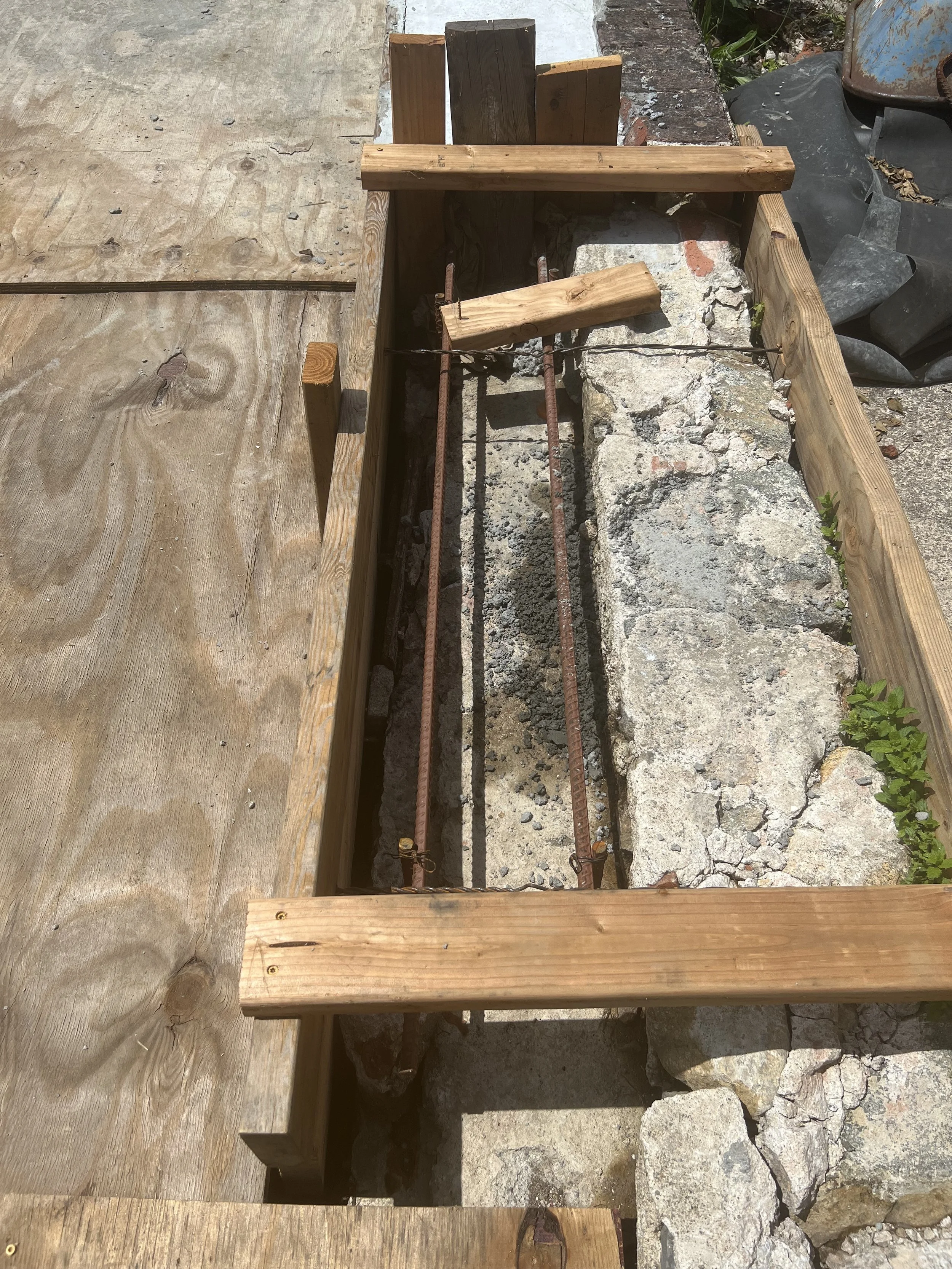 A partially constructed wooden frame with metal reinforcement bars and bricks, indicating ongoing concrete work, outdoors on a sunny day.