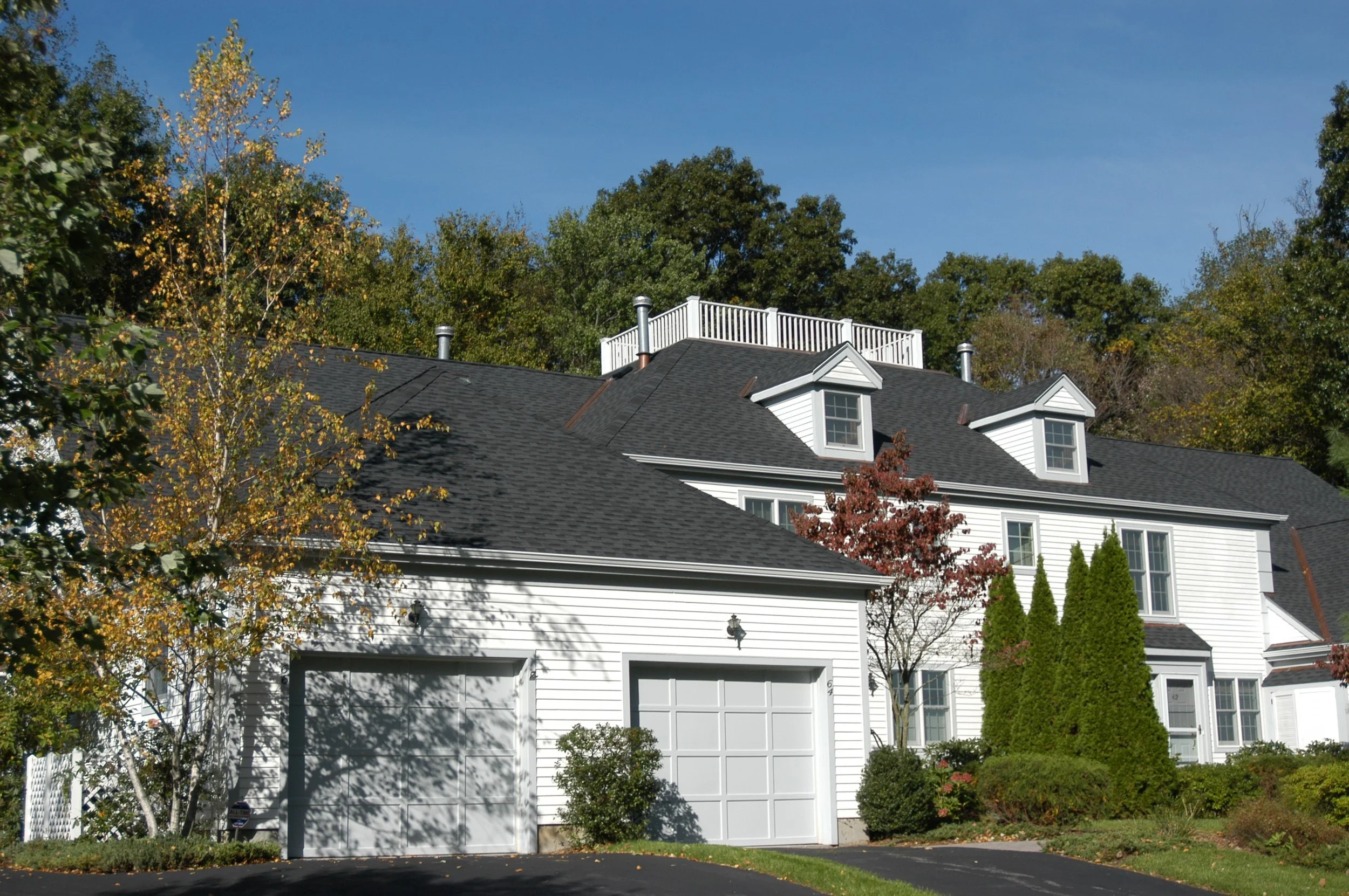 A white house with a black shingle roof and a two-car garage, surrounded by trees with fall foliage, under a clear blue sky.