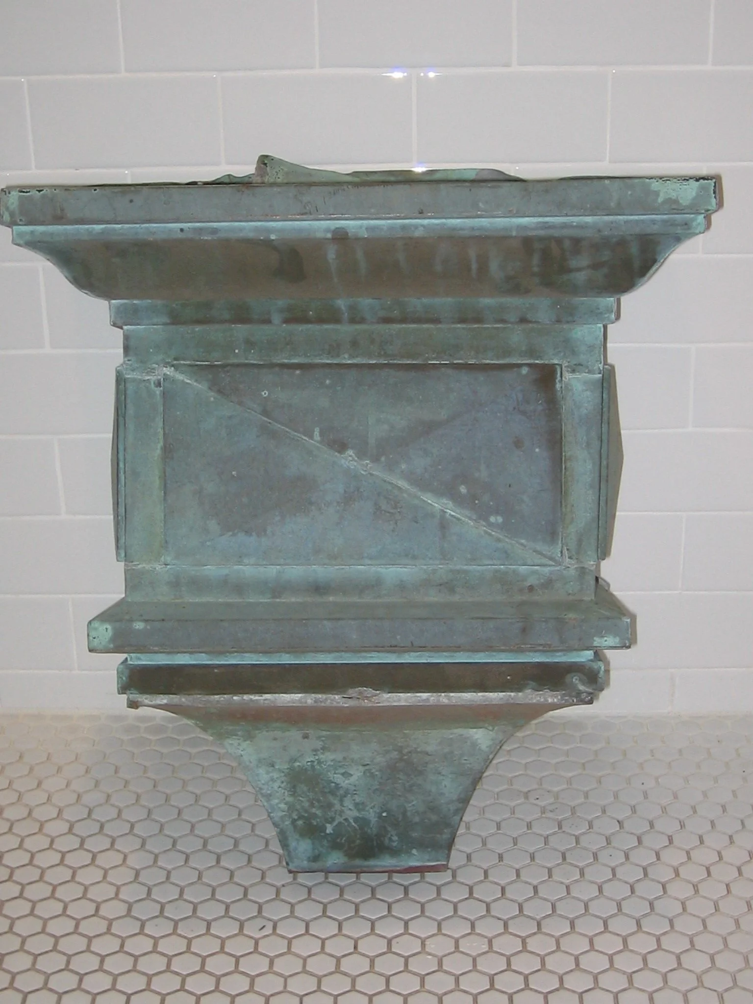 An vintage metal fountain with a basin at the top, mounted on a pedestal, standing on a tiled floor with a white tile wall in the background.