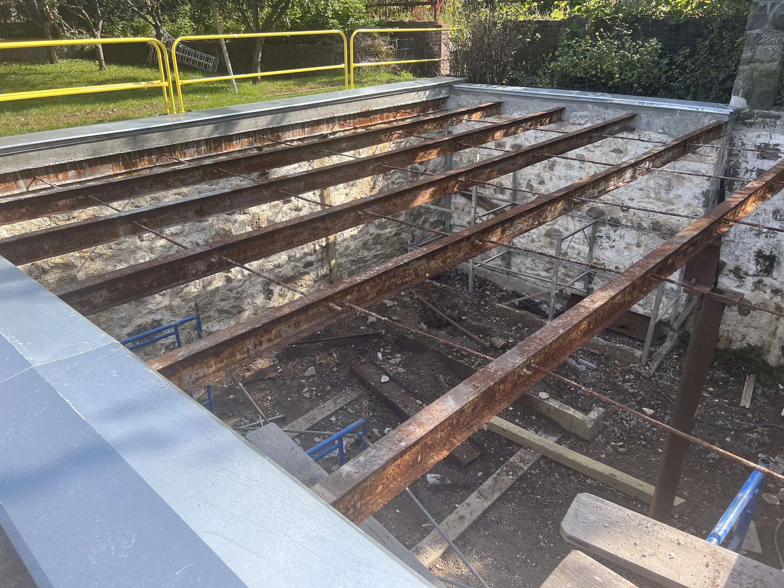 A construction site showing a stone foundation with rusted metal beams and temporary scaffolding, with a yellow safety railing in the background, surrounded by trees and greenery.
