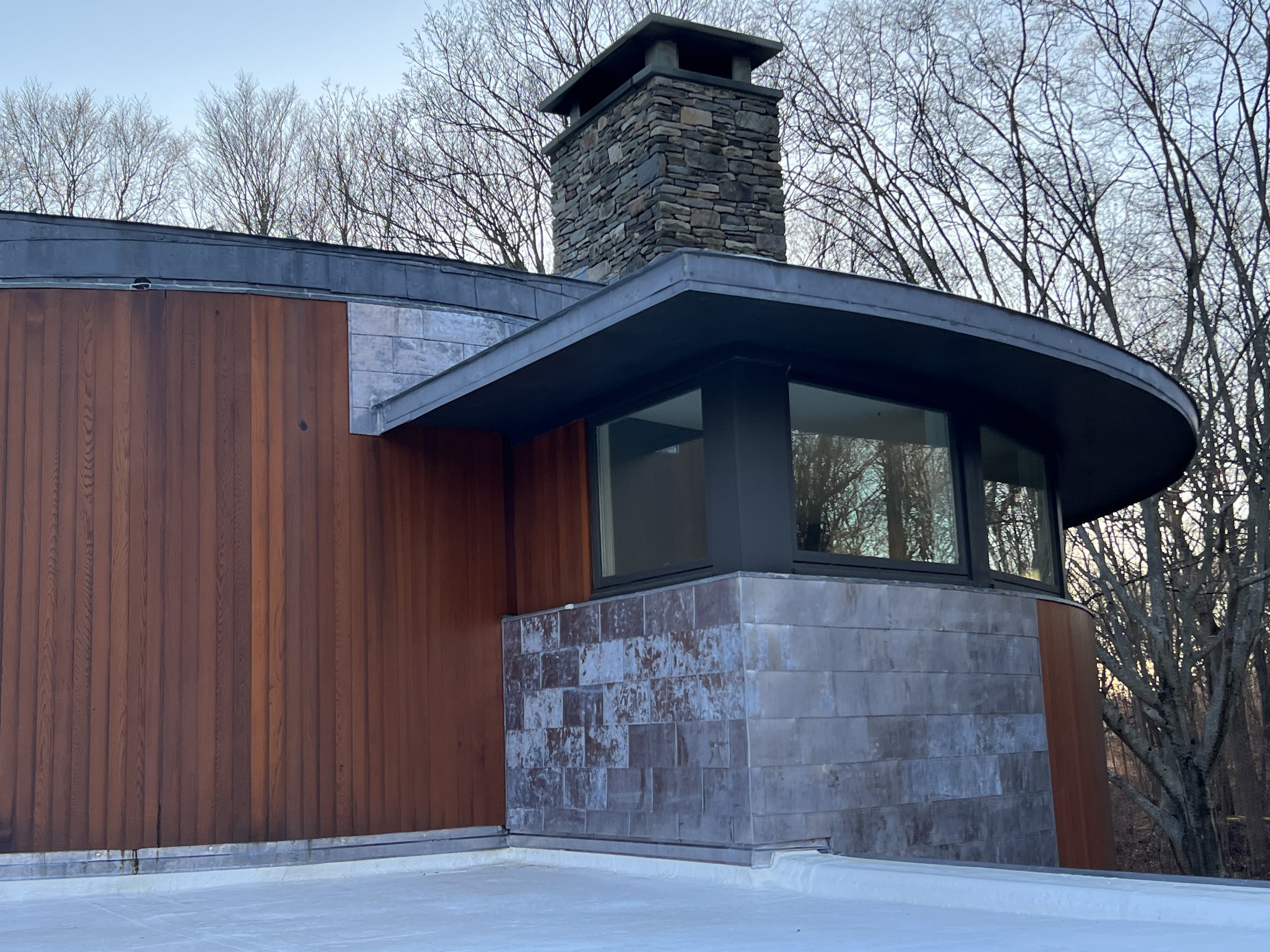 Close-up of a modern house exterior with a curved roof, stone chimney, and mixed wood and stone siding, set against leafless trees in the background.