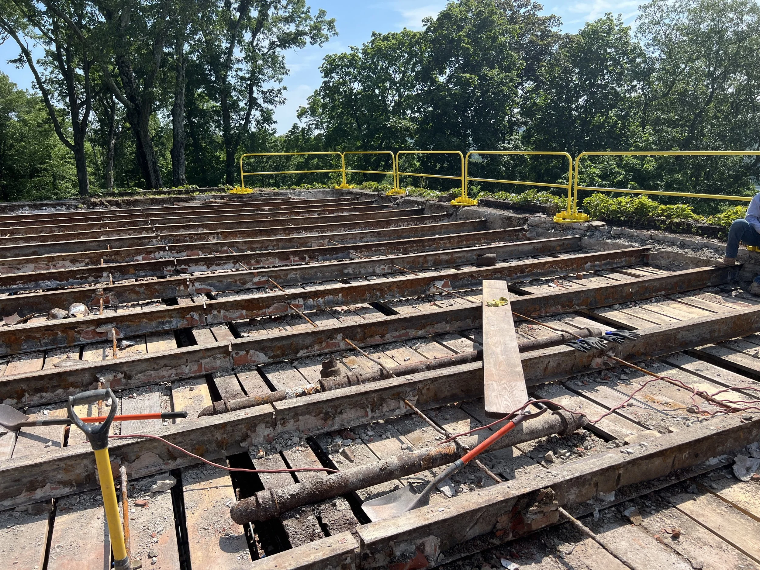 Construction site with stairs under renovation, yellow safety railing, tools, and a person sitting on the right side, surrounded by trees and blue sky.