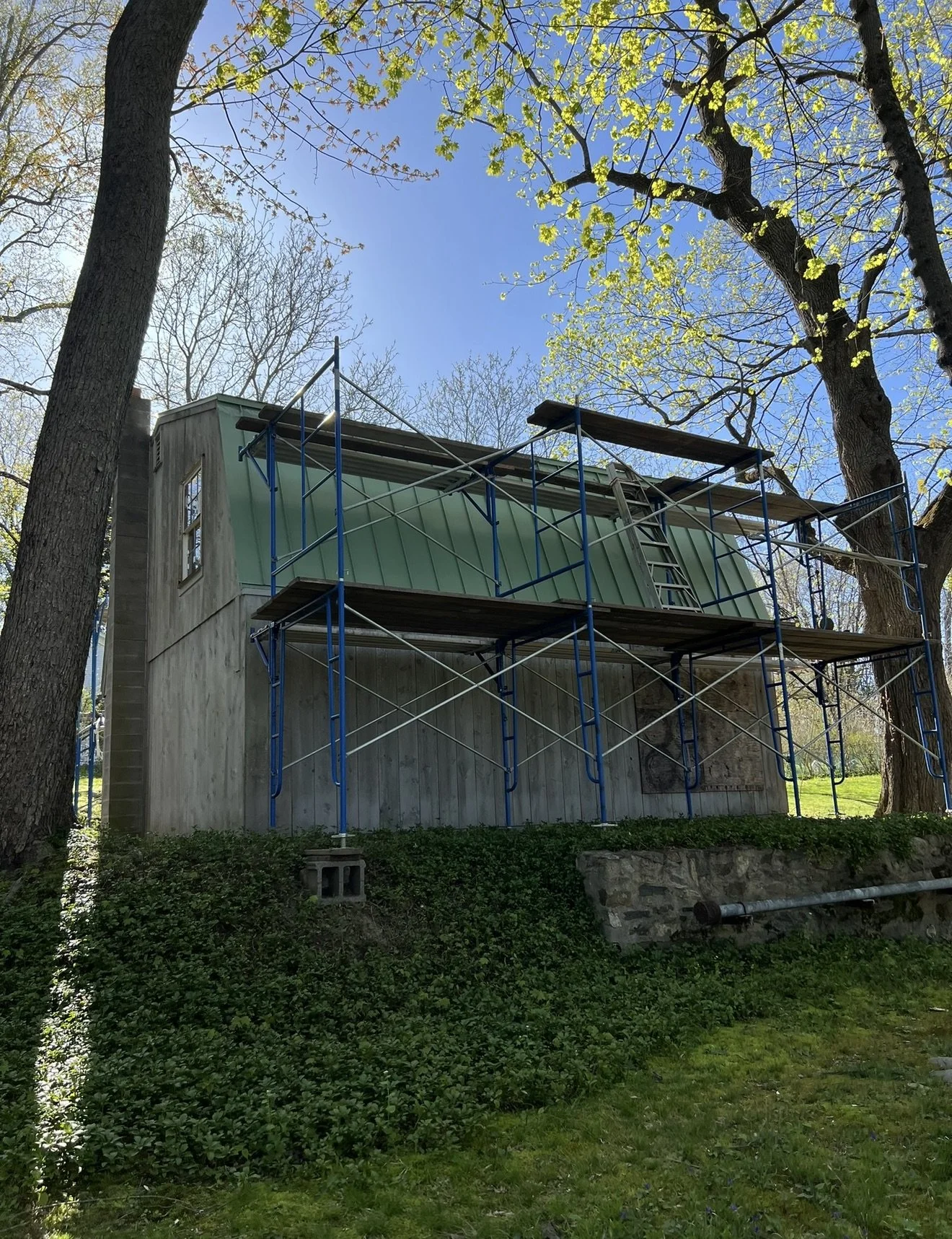 A house under construction with scaffolding around it, situated among trees with green leaves, and a clear blue sky above.