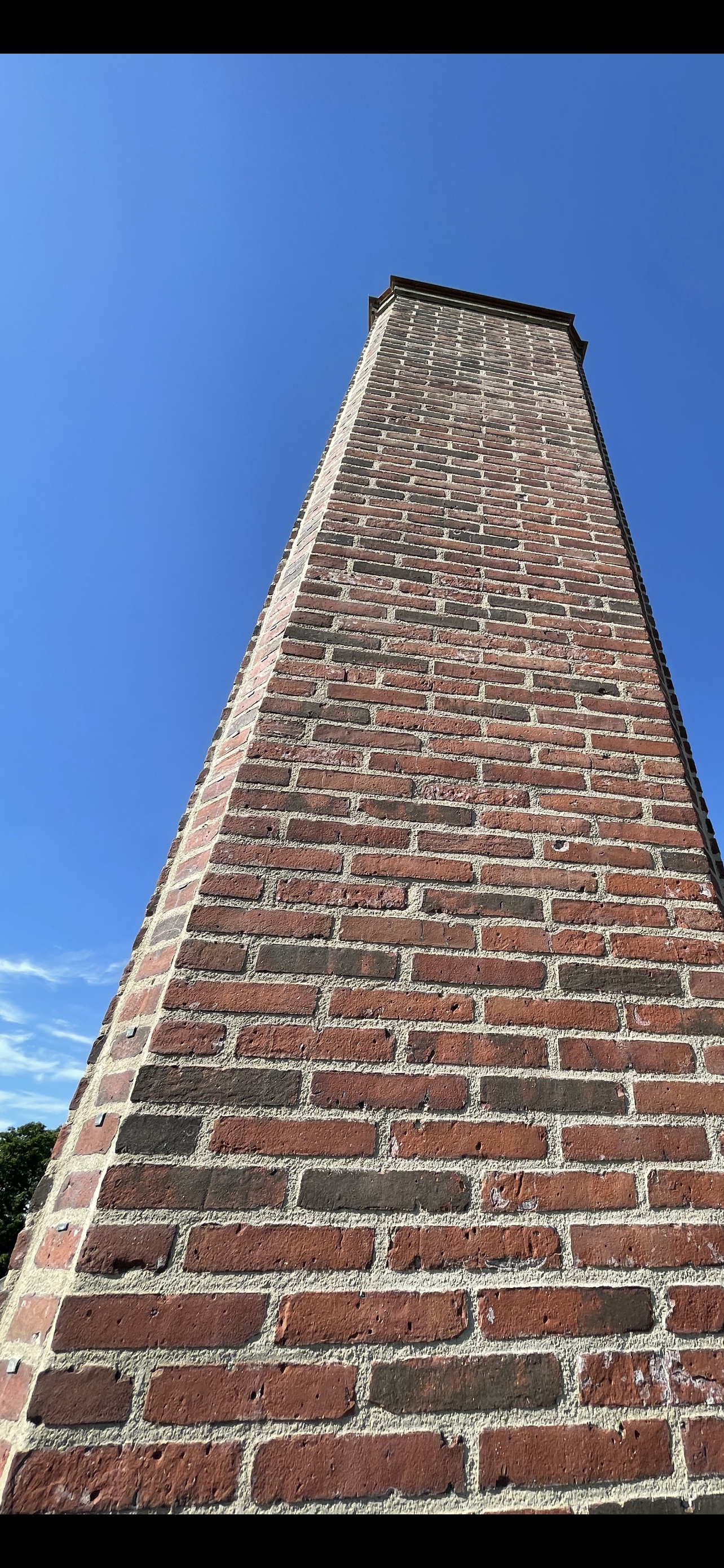 A tall brick chimney seen from below, extending into a clear blue sky, with some green trees at the bottom left corner.