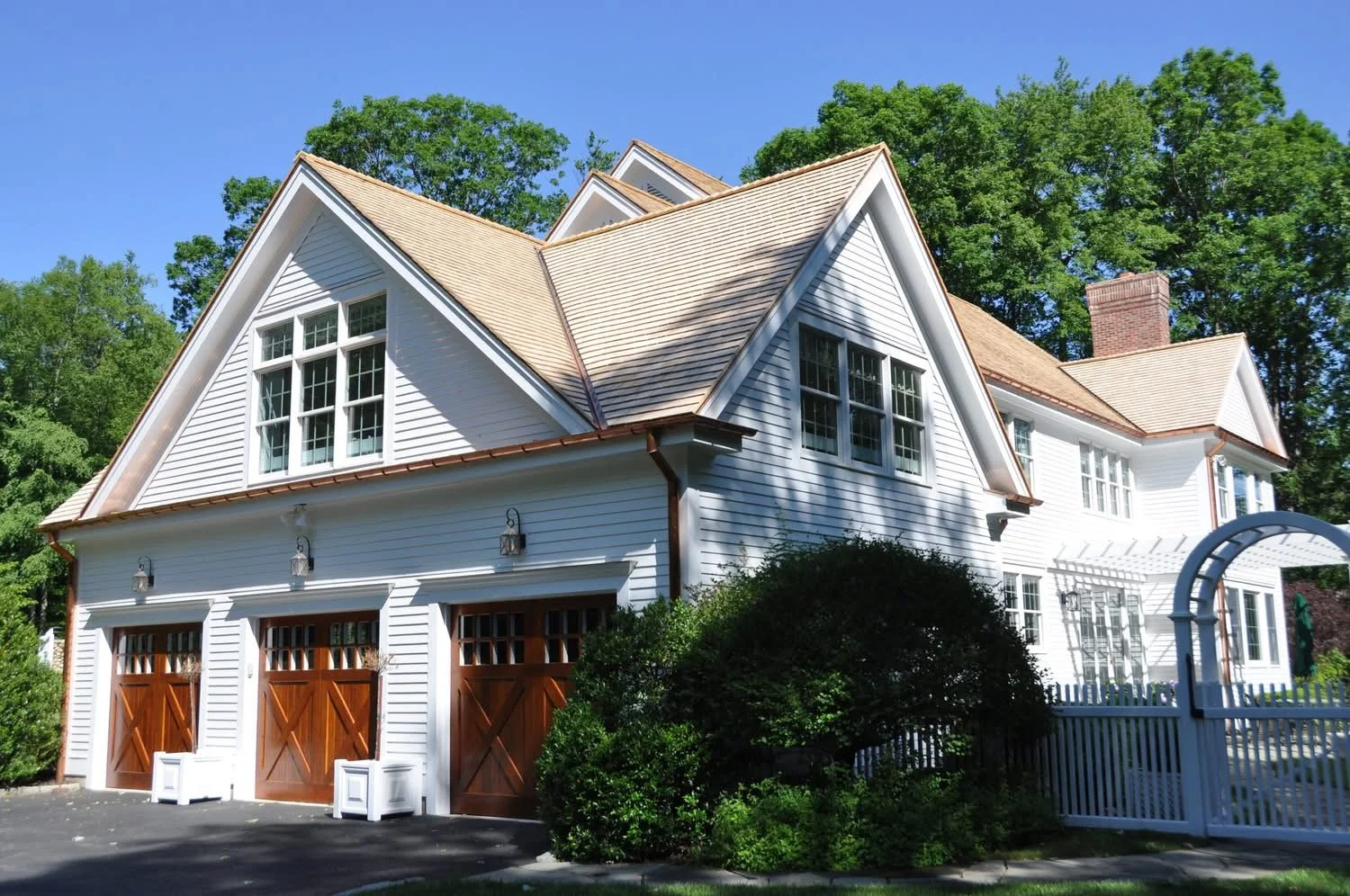 A two-story white house with a steep gable roof, wooden garage doors, multiple windows, surrounded by greenery, and a white picket fence.
