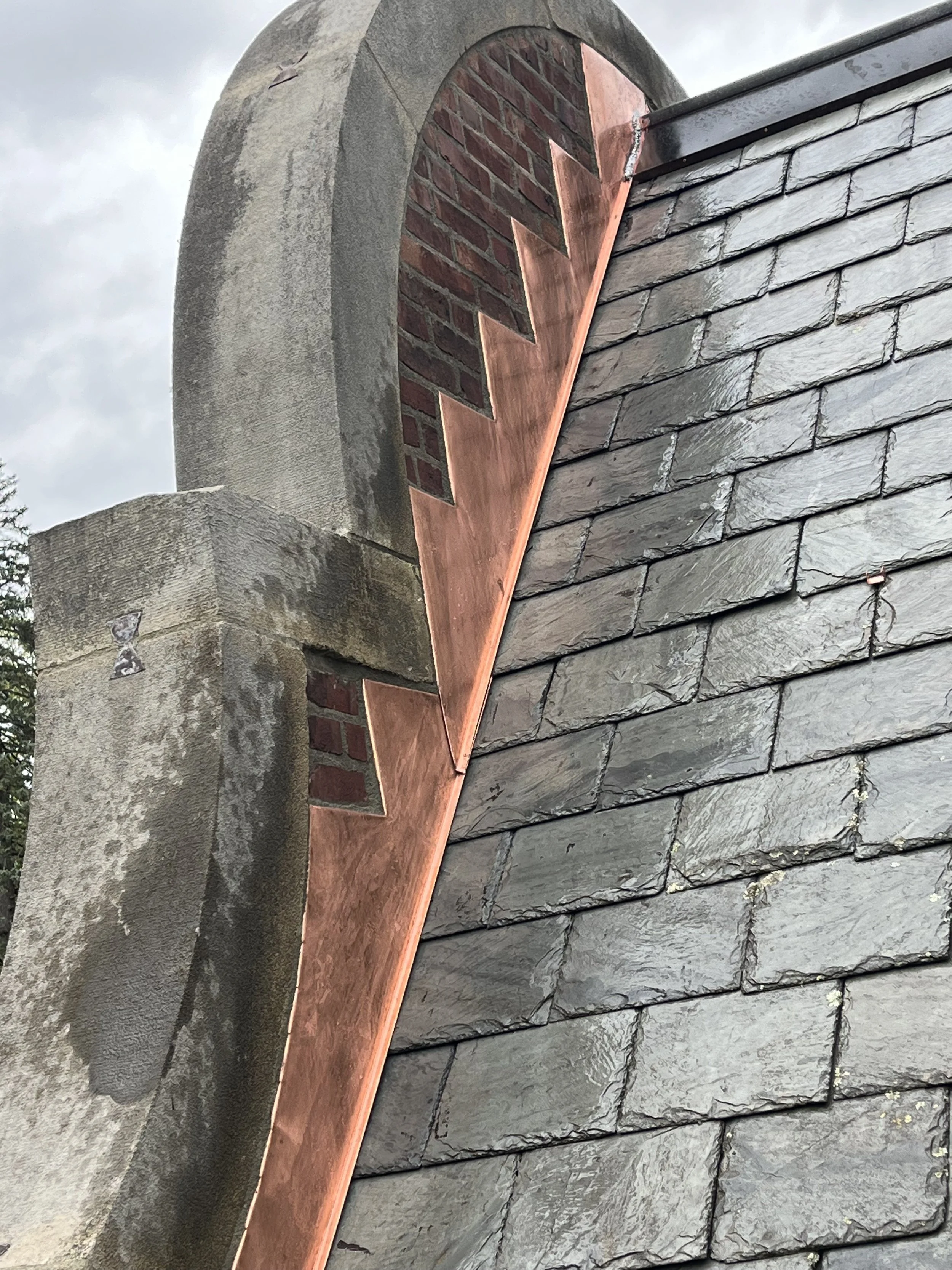 Close-up of a roof with slate shingles, a stone chimney, copper flashing, and a brick arch.