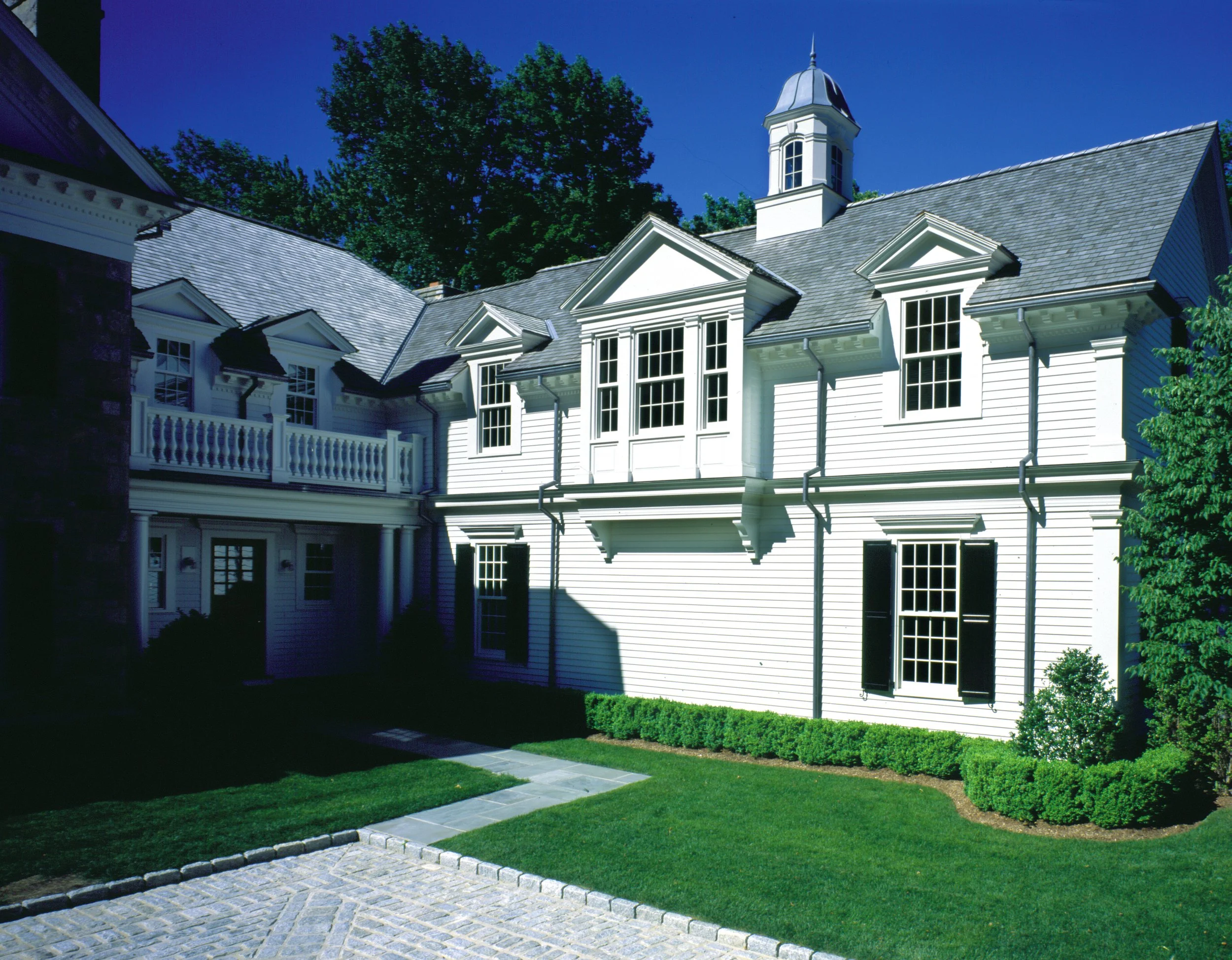 A large white traditional-style house with multiple gabled roofs, a small tower with a spire, tall windows, and a porch. The house is surrounded by well-maintained green lawn, bushes, and trees, with a sidewalk in front.