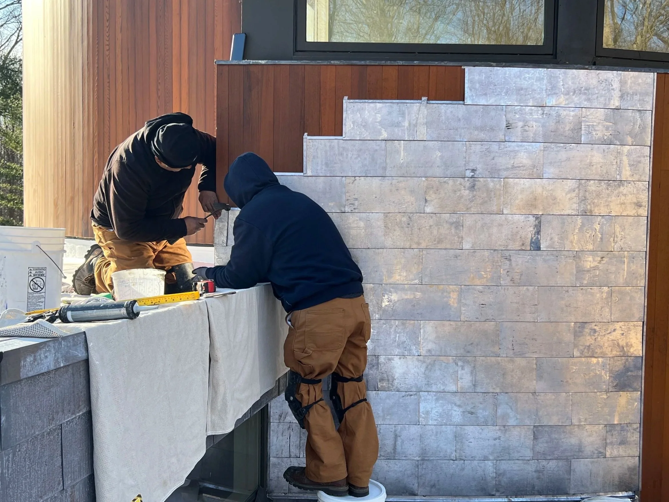 Two workers install stone tiles on an exterior wall, with one kneeling and using a tool, and the other standing in front, both wearing work pants and jackets, on a balcony or terrace with a wooden railing and glass door.