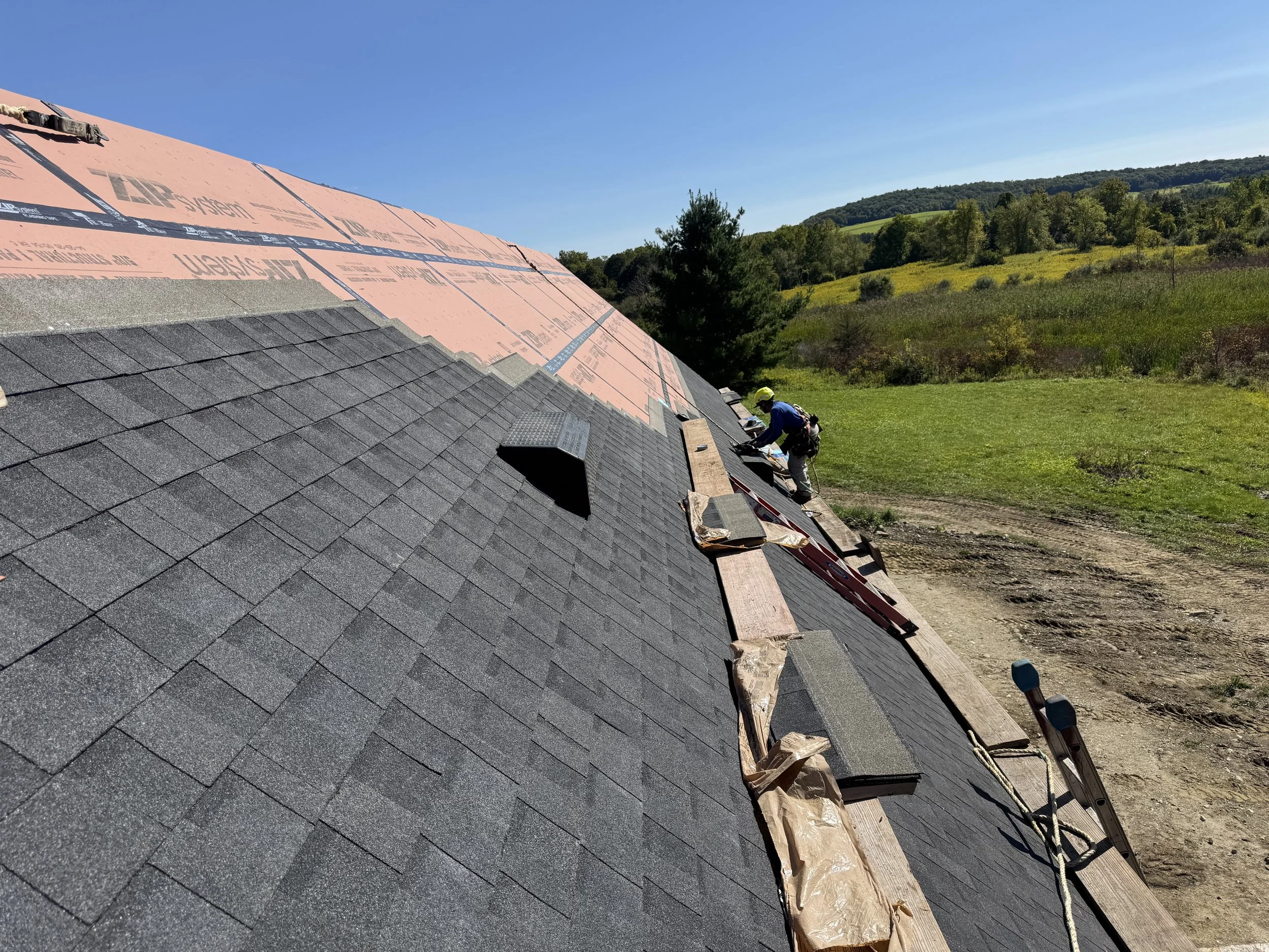 Roof undergoing repair with workers installing asphalt shingles in a rural area with green hills and clear blue sky in the background.