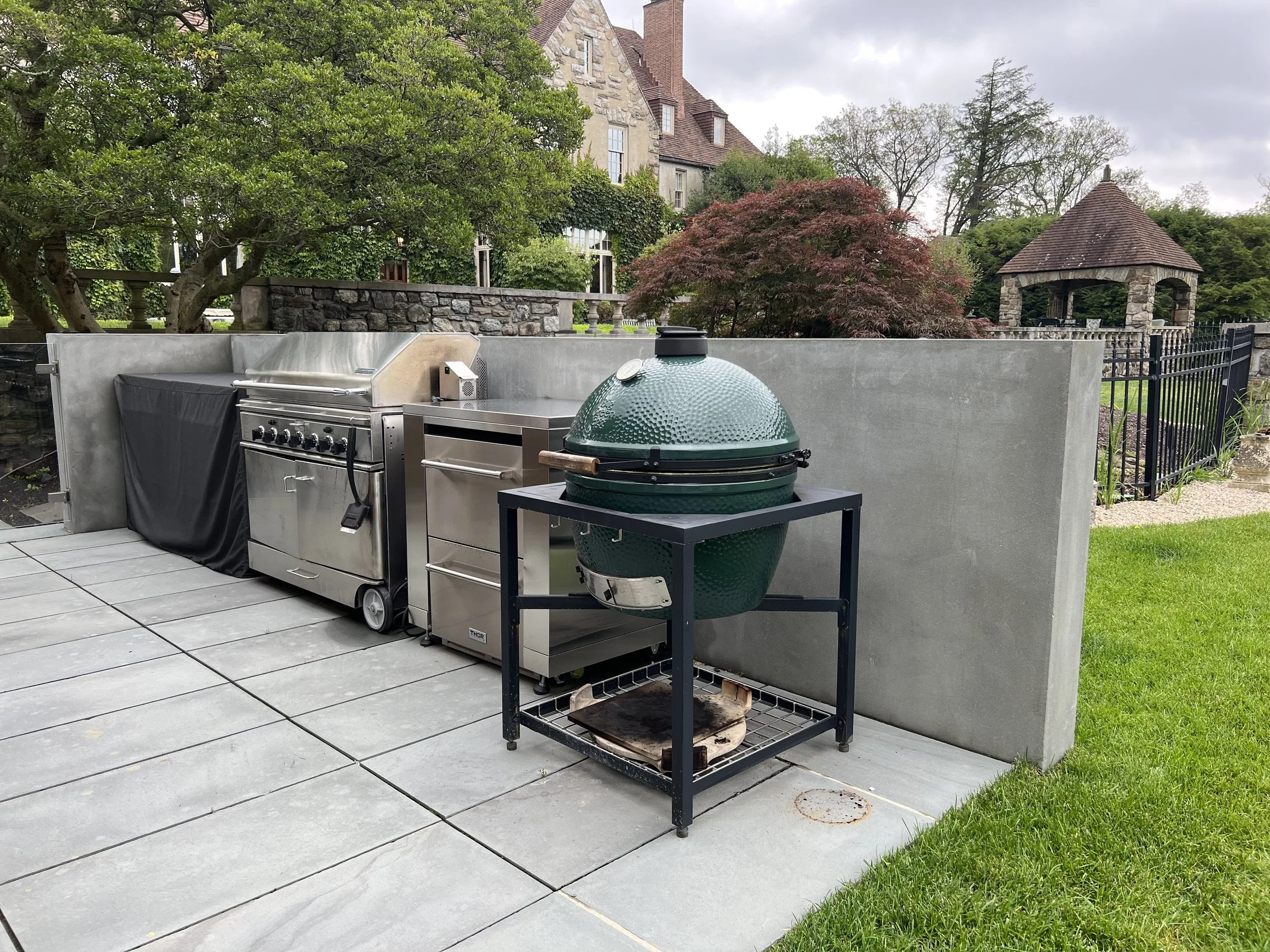 Outdoor patio with stainless steel grill and a green ceramic Kamado-style barbecue, surrounded by a concrete wall, with a stone house and lush trees in the background, under a cloudy sky.