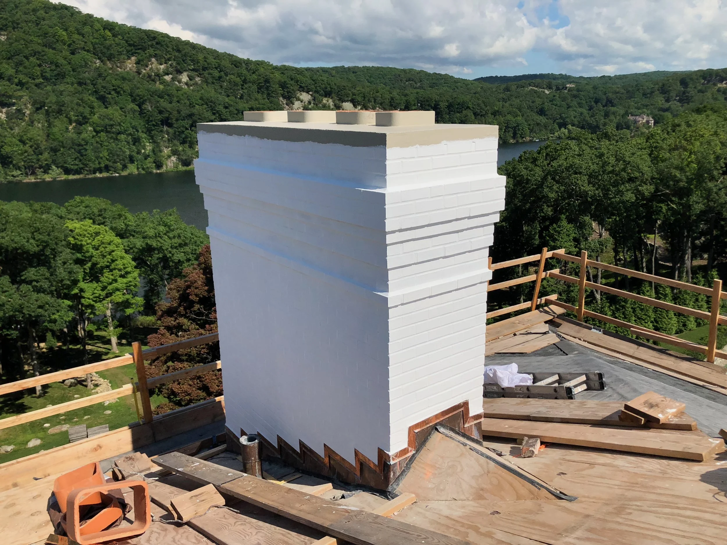 A newly constructed white brick chimney on a rooftop with protective wooden railings around the edge, overlooking a lush green landscape with trees, hills, and a body of water in the distance under a partly cloudy sky.