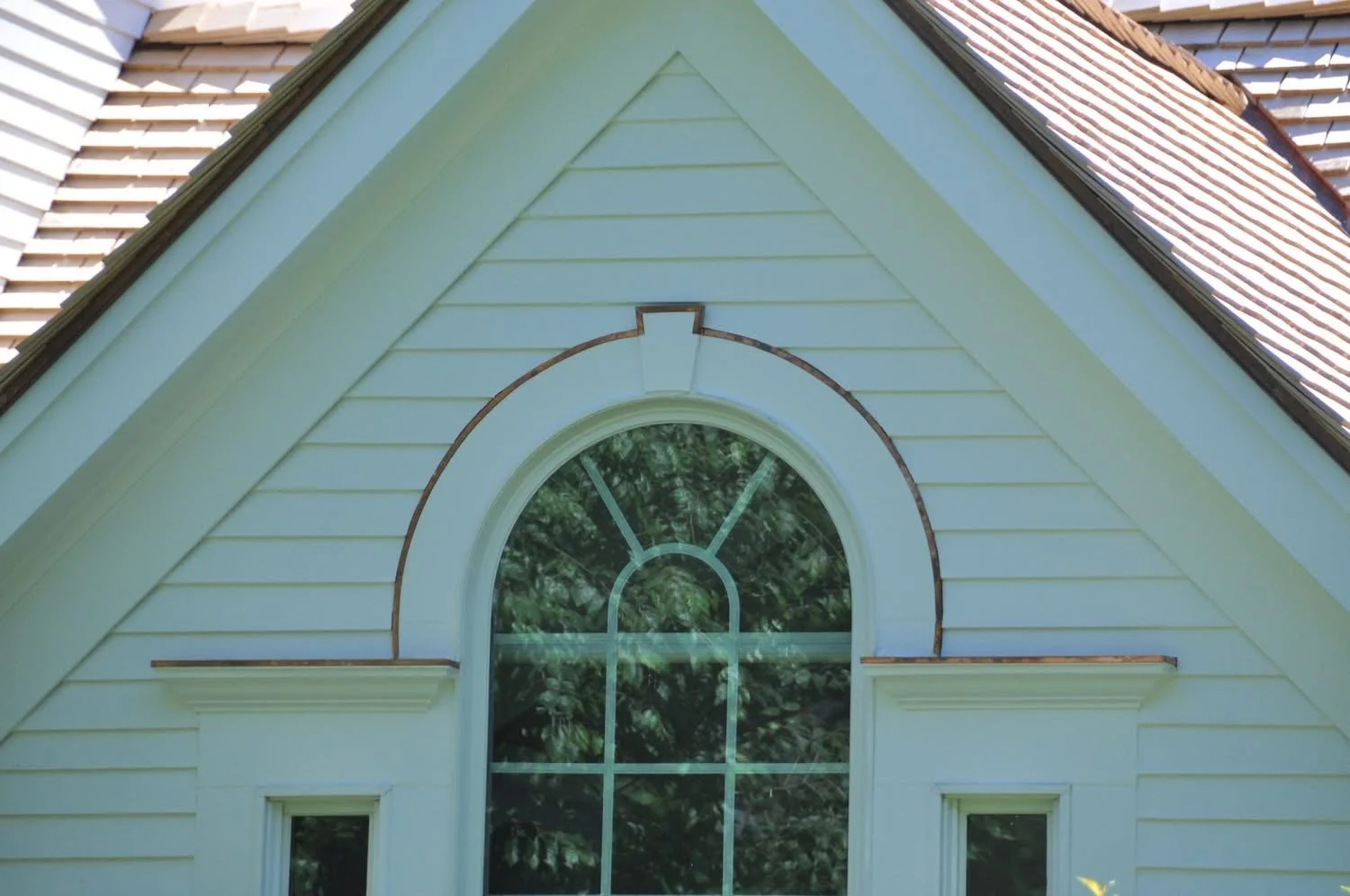 Close-up of a house with a pointed roof, white horizontal siding, and a large arched window with decorative grid patterns, surrounded by trees reflected in the glass.