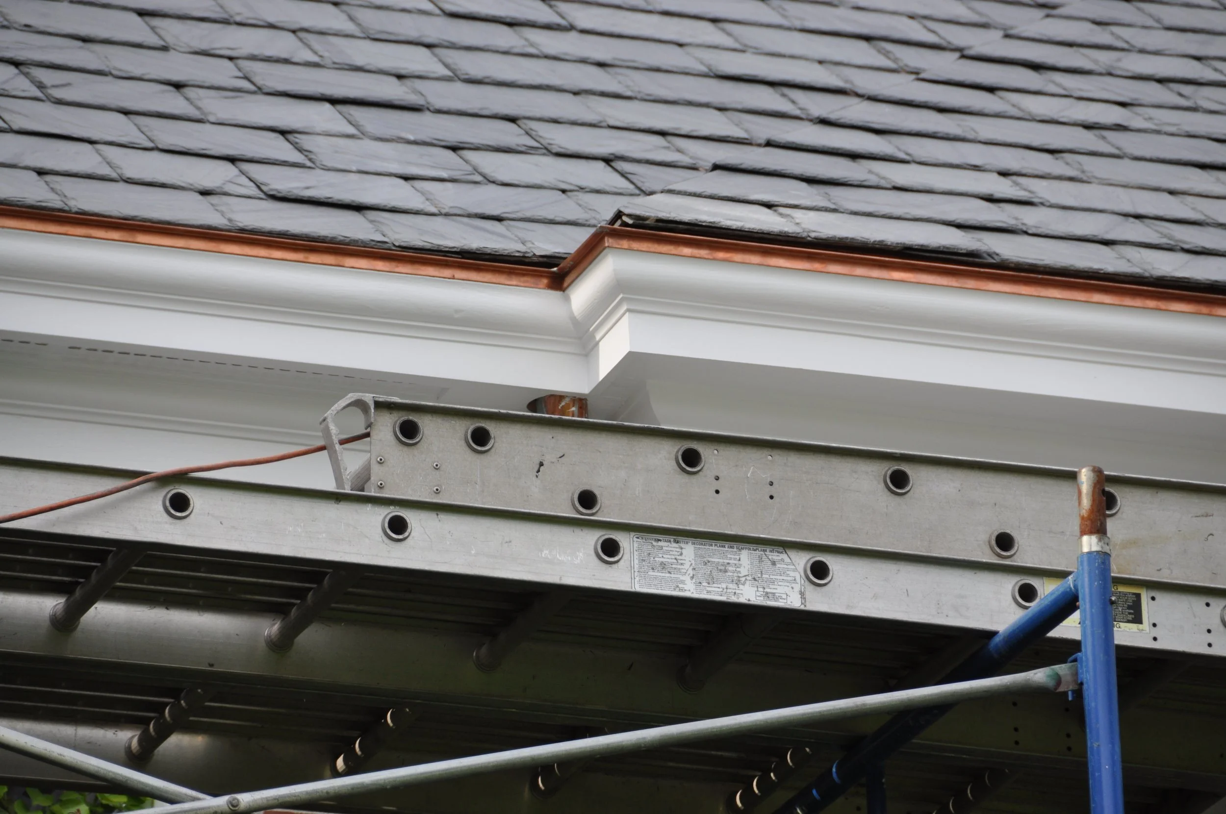 Close-up of a construction scaffolding at the edge of a building with grey slate roof shingles and white trim.