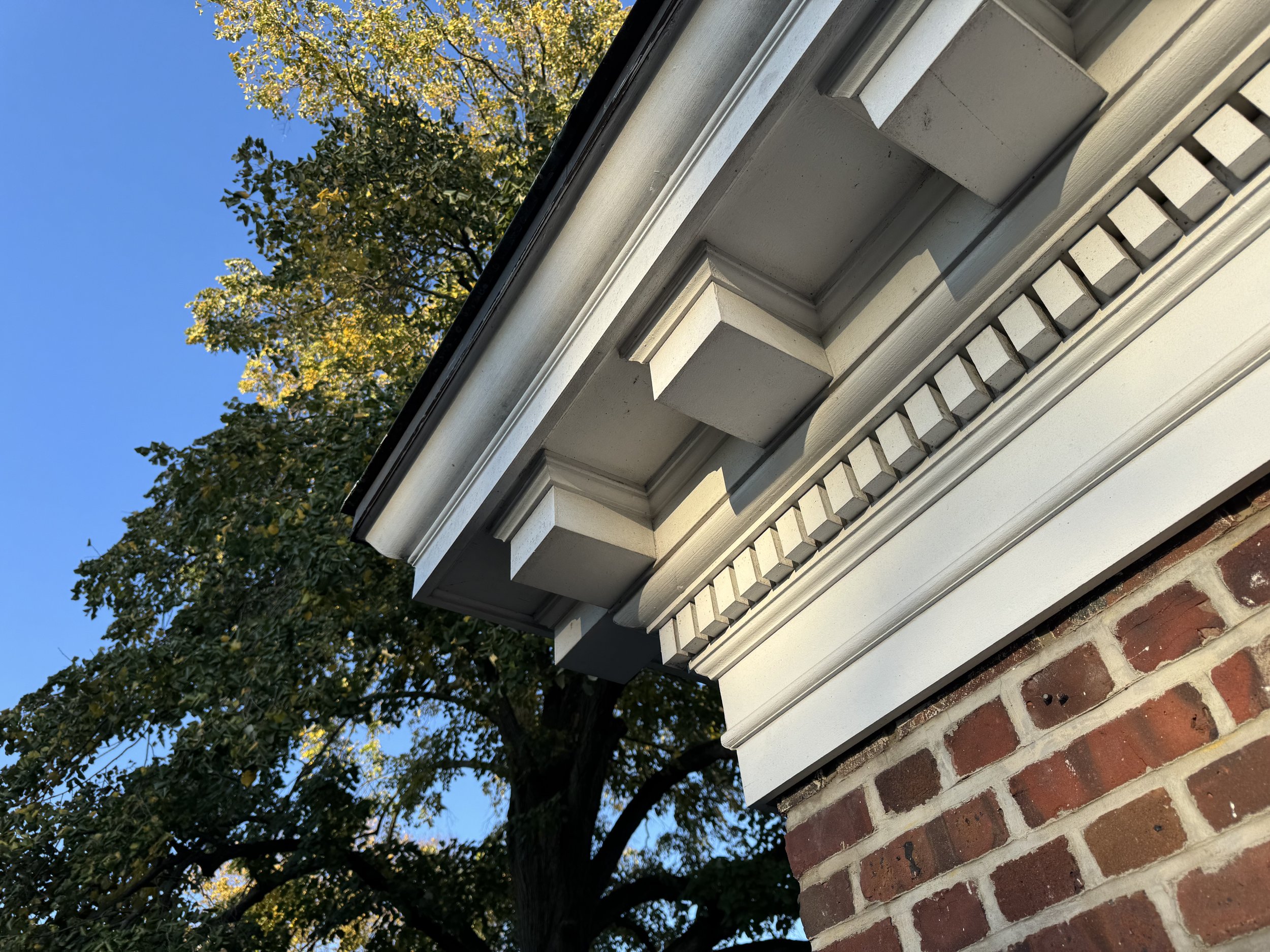 Close-up of architectural details on a house's exterior, including decorative molding and a brick foundation, with a background of a blue sky and trees with green and yellow leaves.