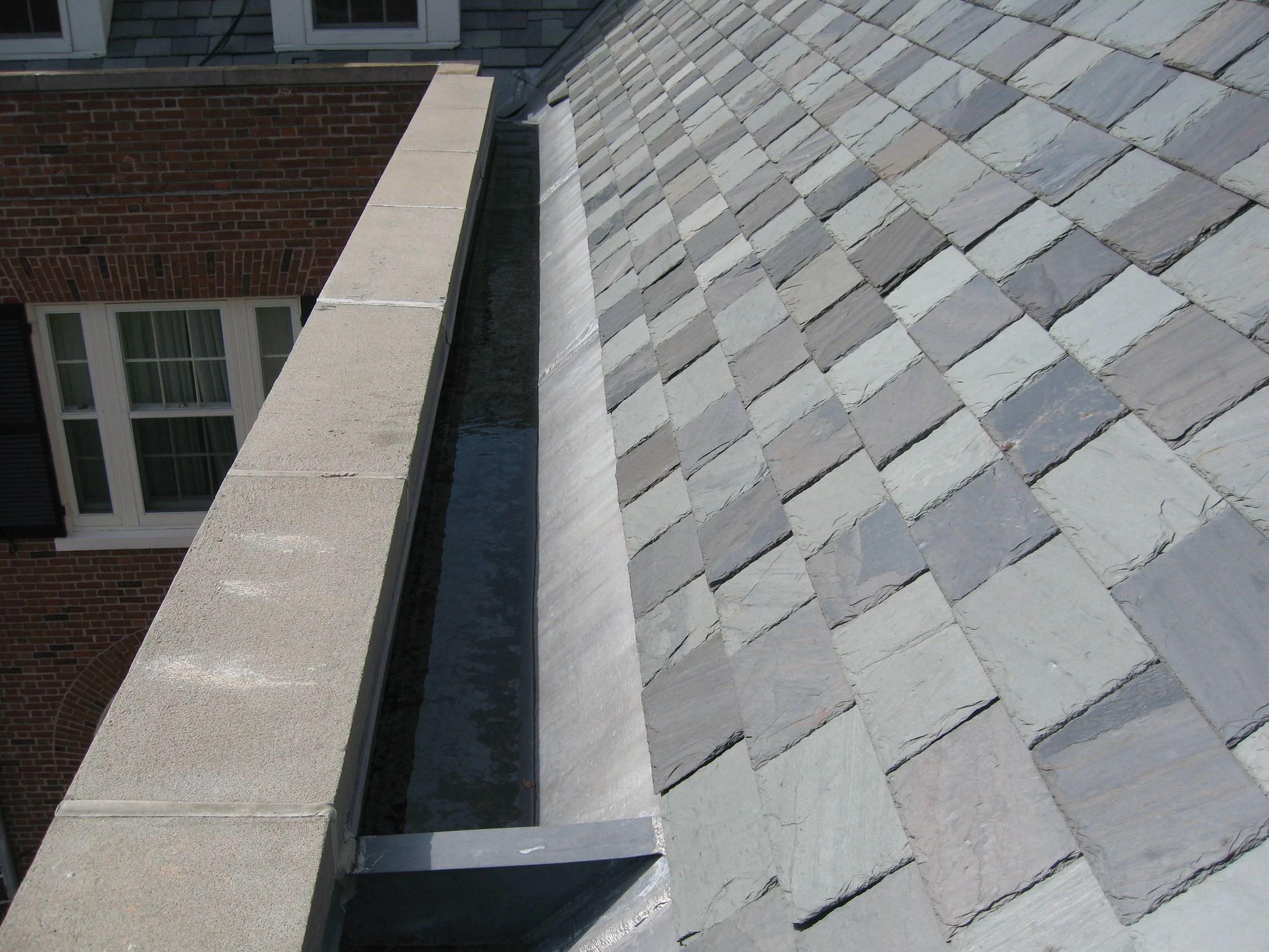 A rooftop with gray slate shingles, a brick building with white-framed windows, and a concrete parapet wall.