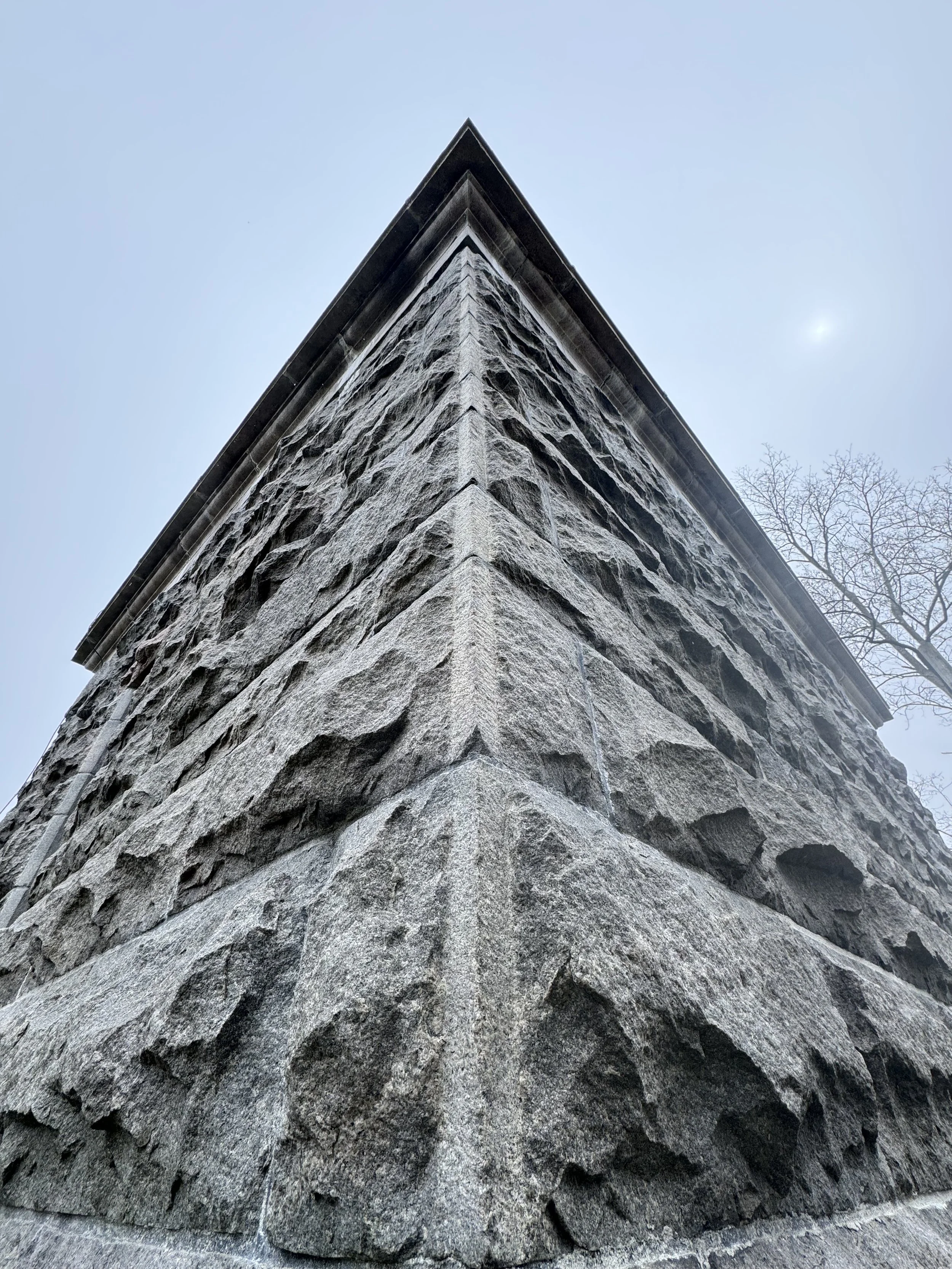 Close-up of a stone building corner with textured gray stones and a dark overhanging roof, under a clear sky with leafless trees in the background.