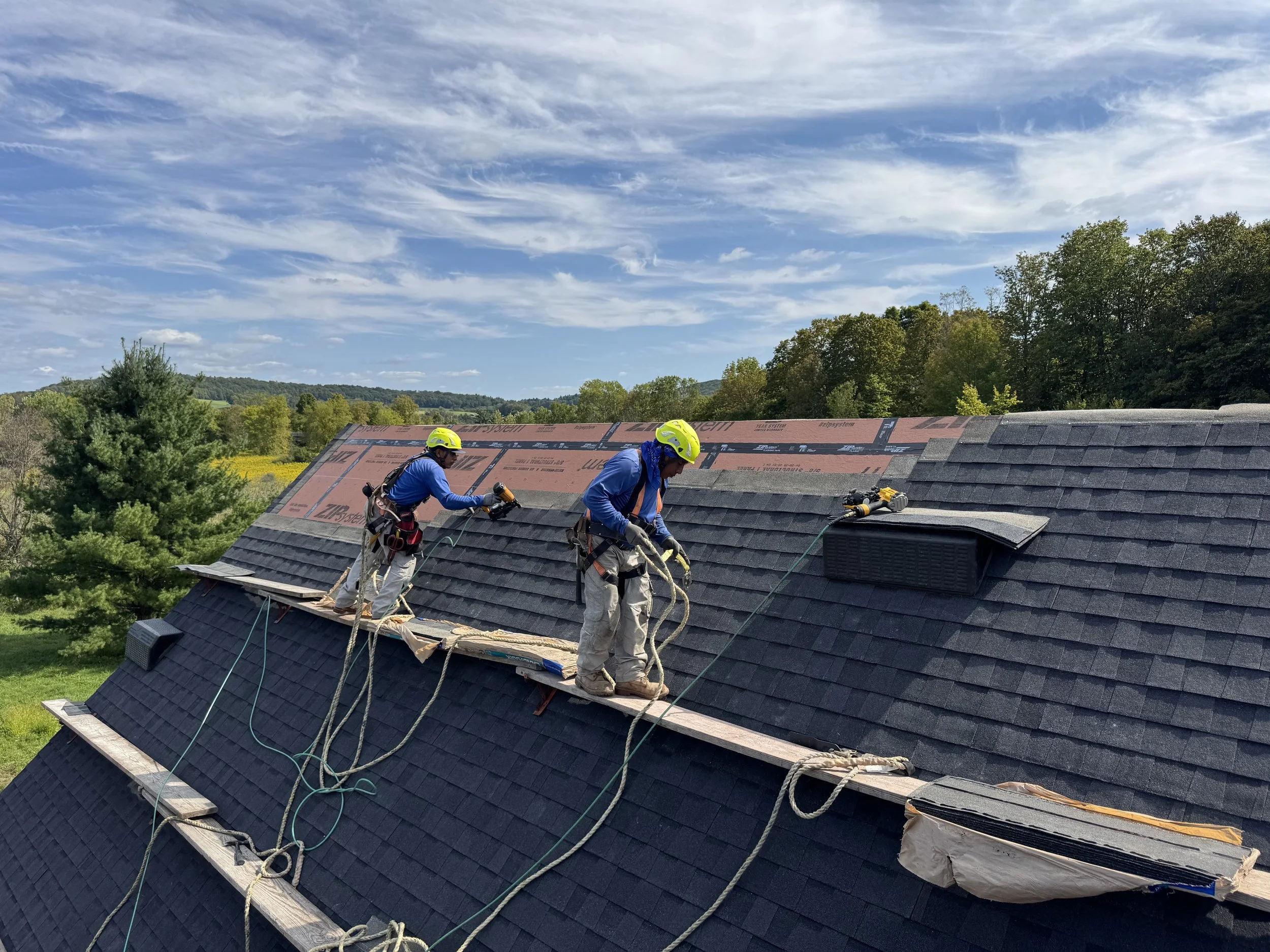 Two workers wearing yellow safety helmets repairing or installing roof shingles on a sloped roof, with tools and safety ropes.