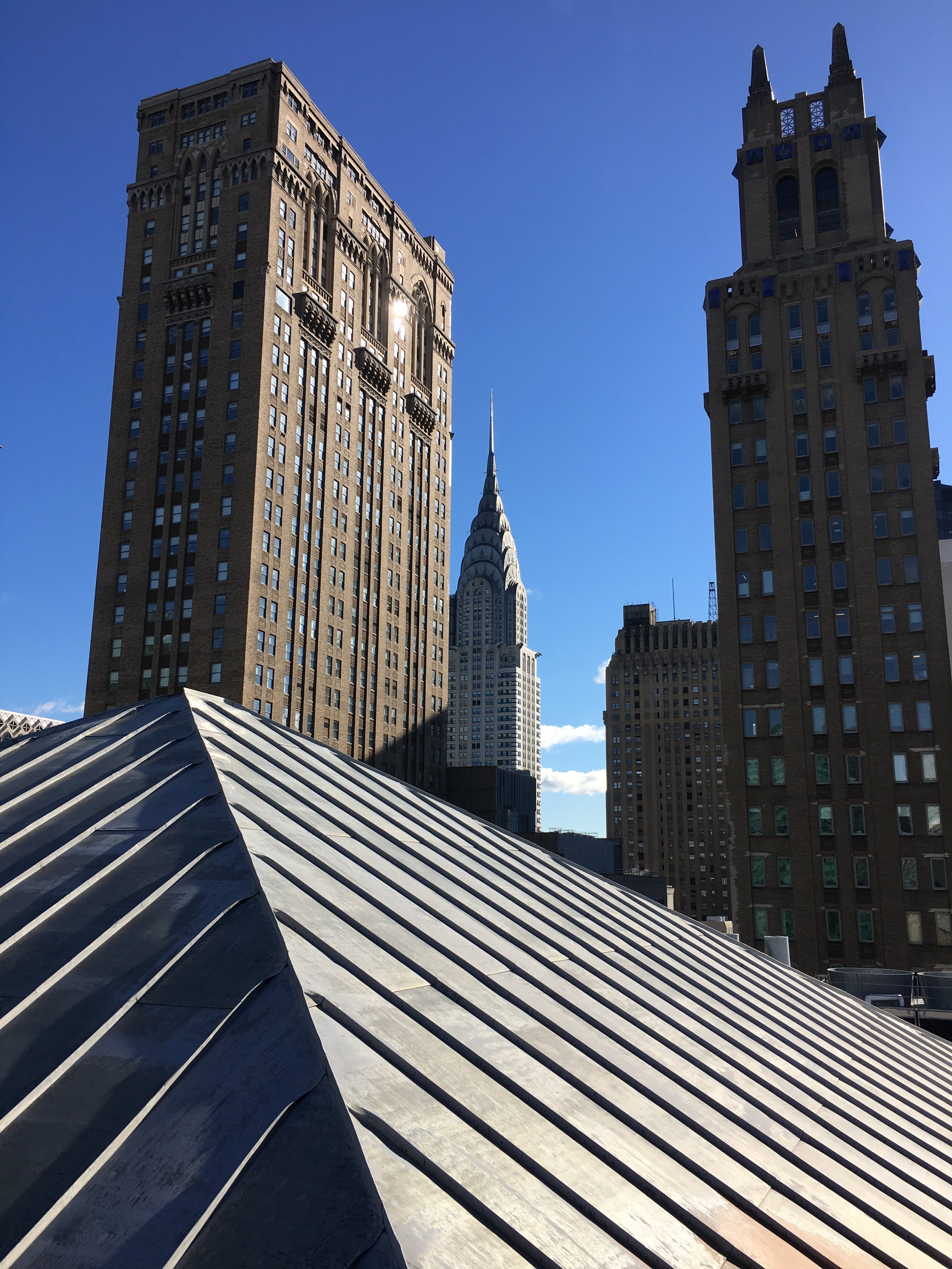 City skyline with tall buildings, including the Chrysler Building, under a clear blue sky. The foreground features a metal rooftop with slanted panels.