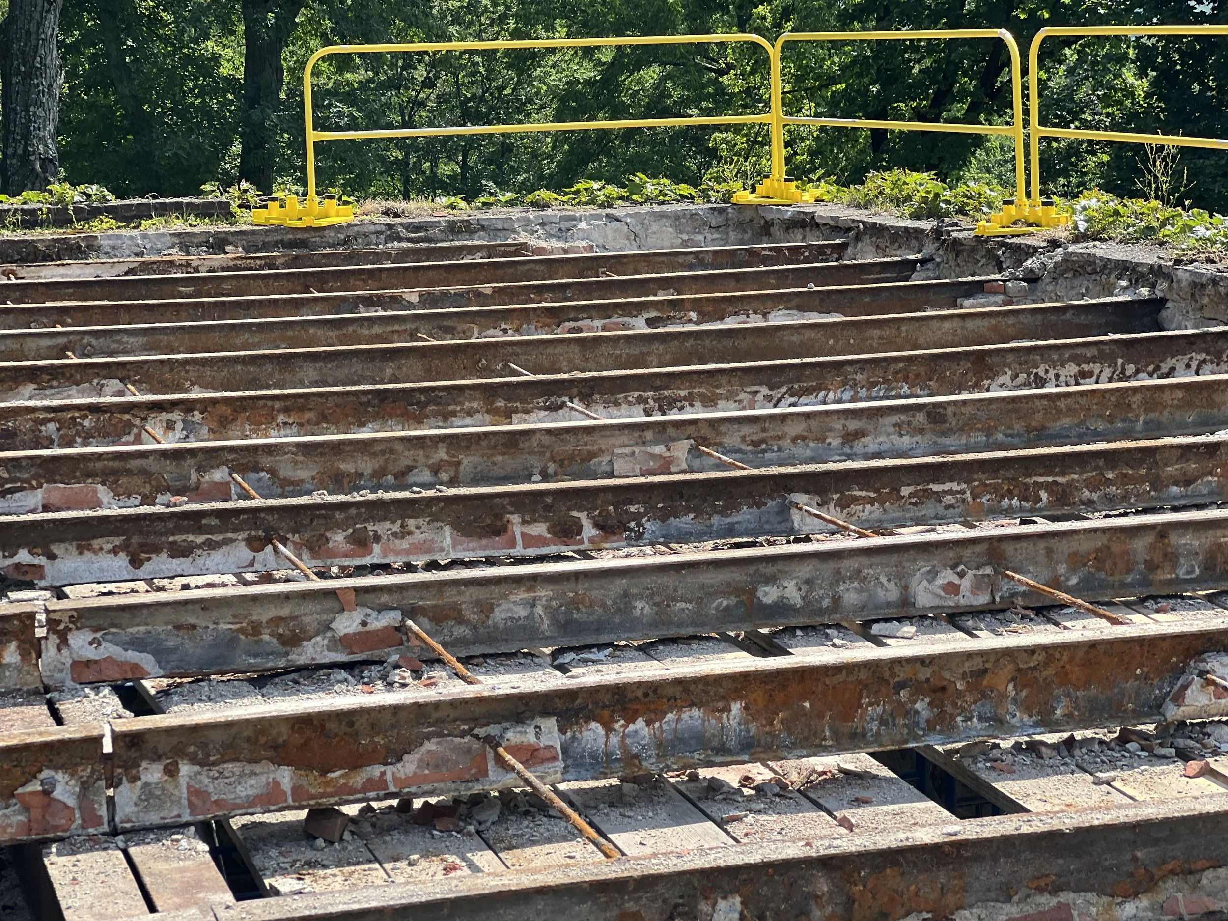 Bricks and concrete debris on rusted metal stairs, with yellow safety barriers at the top, surrounded by green trees.