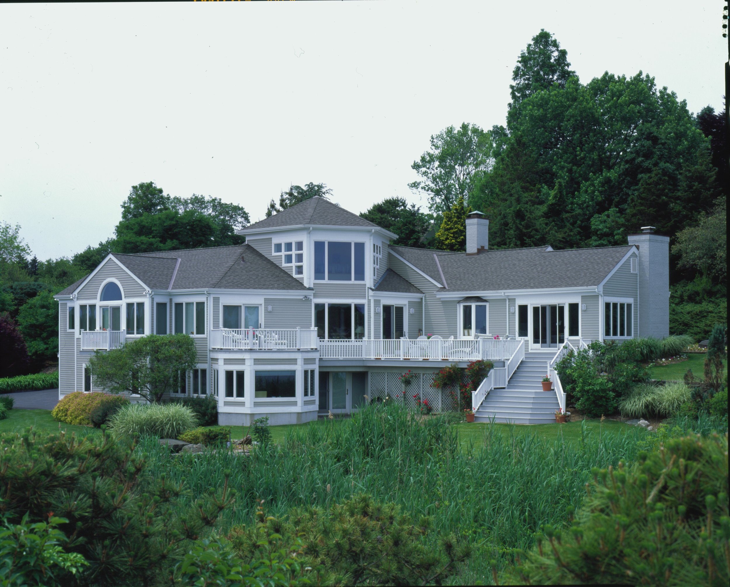 A large, modern house with multiple stories, large windows, and a spacious deck, surrounded by lush green trees and bushes.