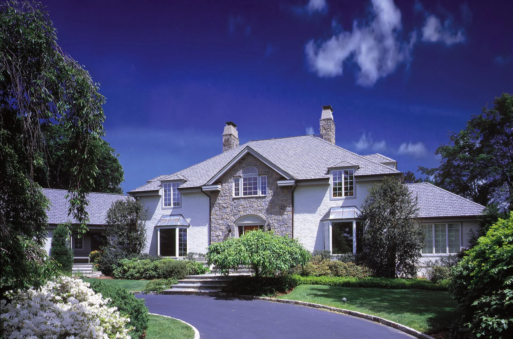 Large house with stone and white exterior, multiple chimneys, surrounded by lush green trees and well-manicured garden under a dark blue sky.