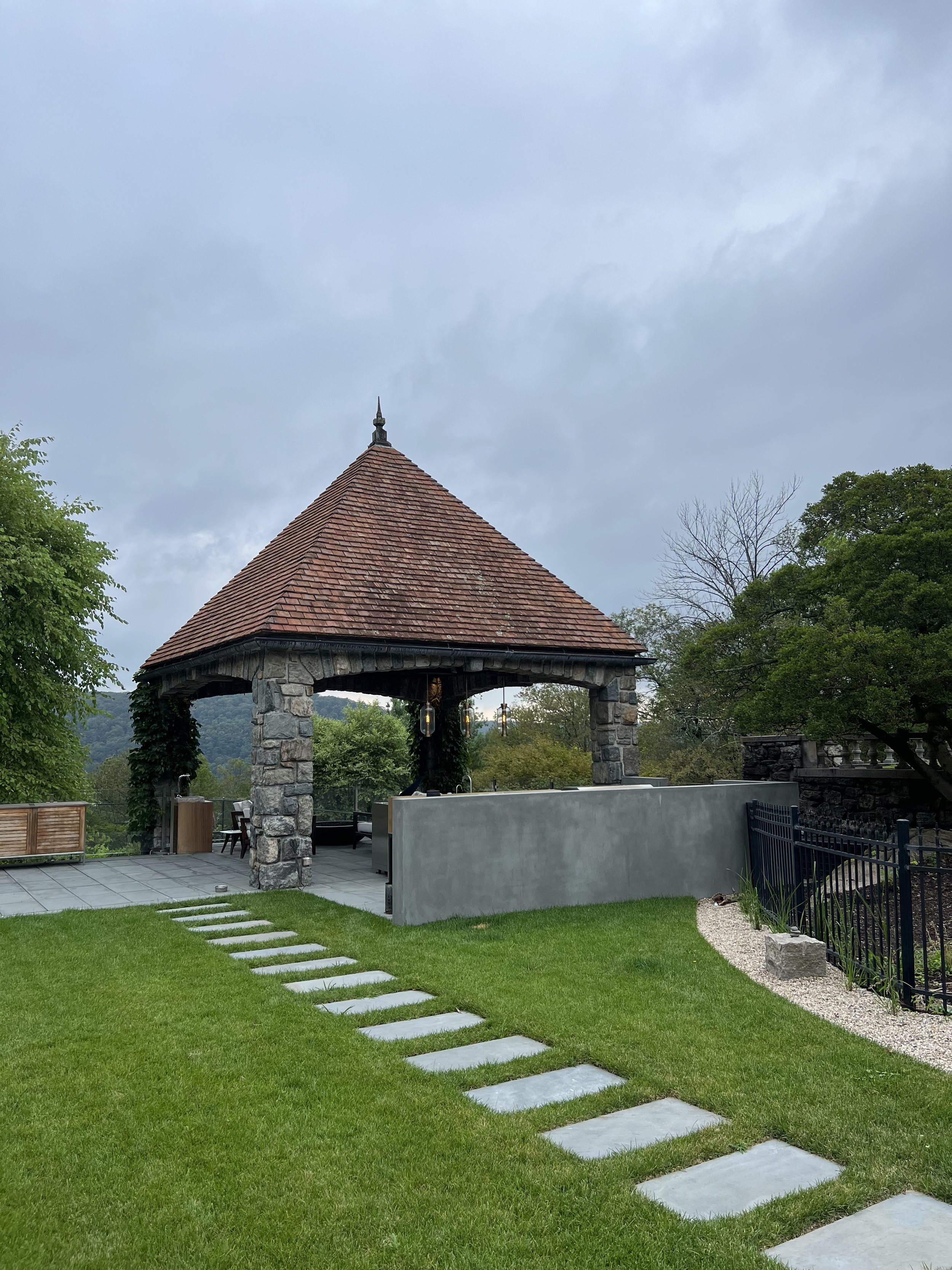 A small stone and wood pavilion with a red tiled roof and hanging lanterns, surrounded by a green lawn with a stepping stone path, trees, and a cloudy sky.