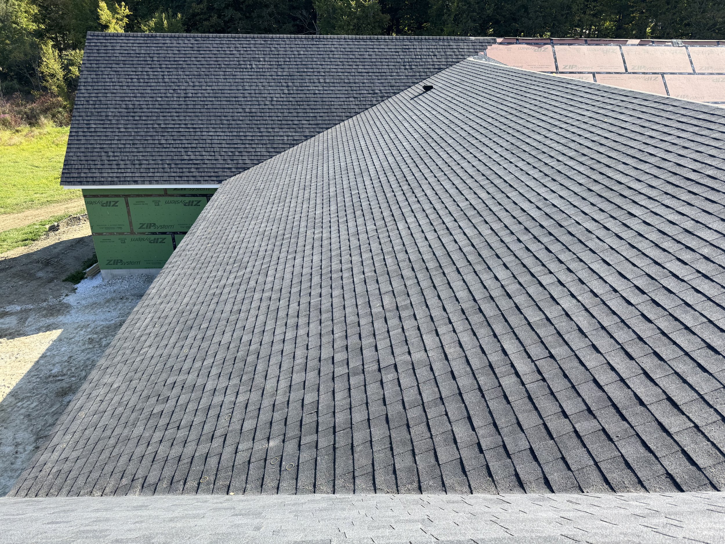 A house roof with new asphalt shingles, part of the roof under construction with exposed roofing material, and surrounded by trees and grass.