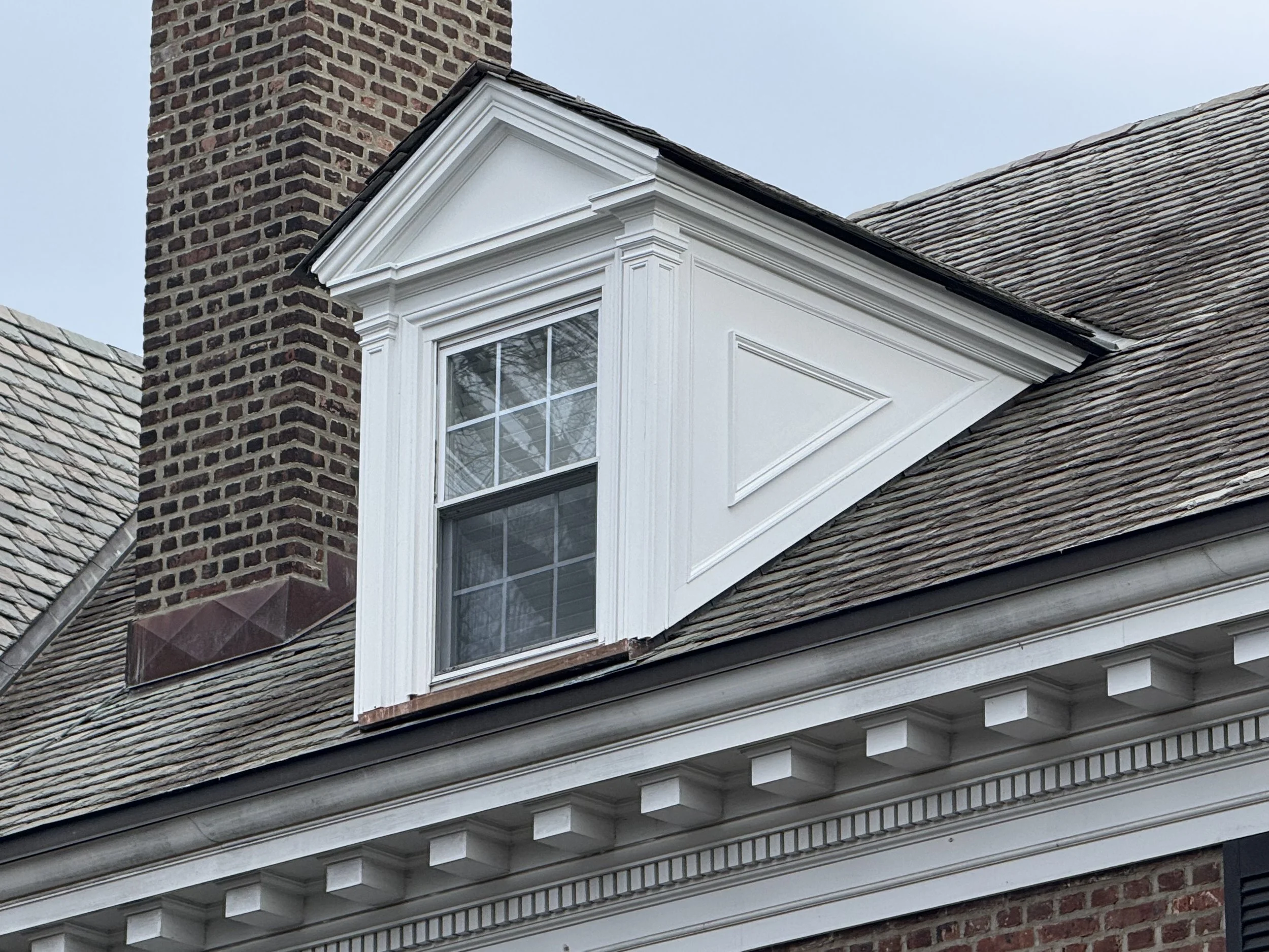 Close-up of a house's roof and chimney, featuring a dormer window with white trim and a roof covered in shingles.