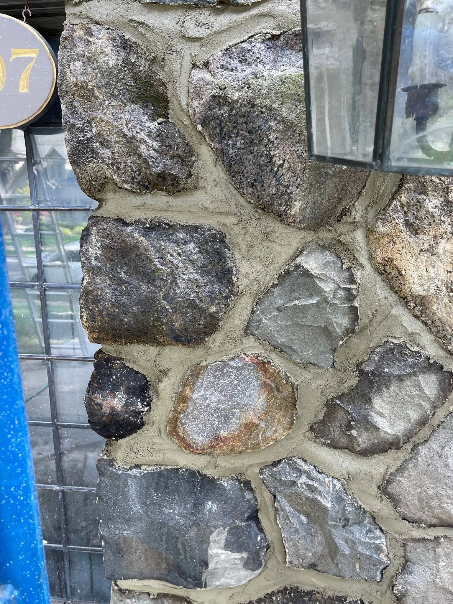 Close-up of a stone wall with various large, irregular-shaped rocks embedded in mortar, with a glass panel or lantern partially visible on the right side.