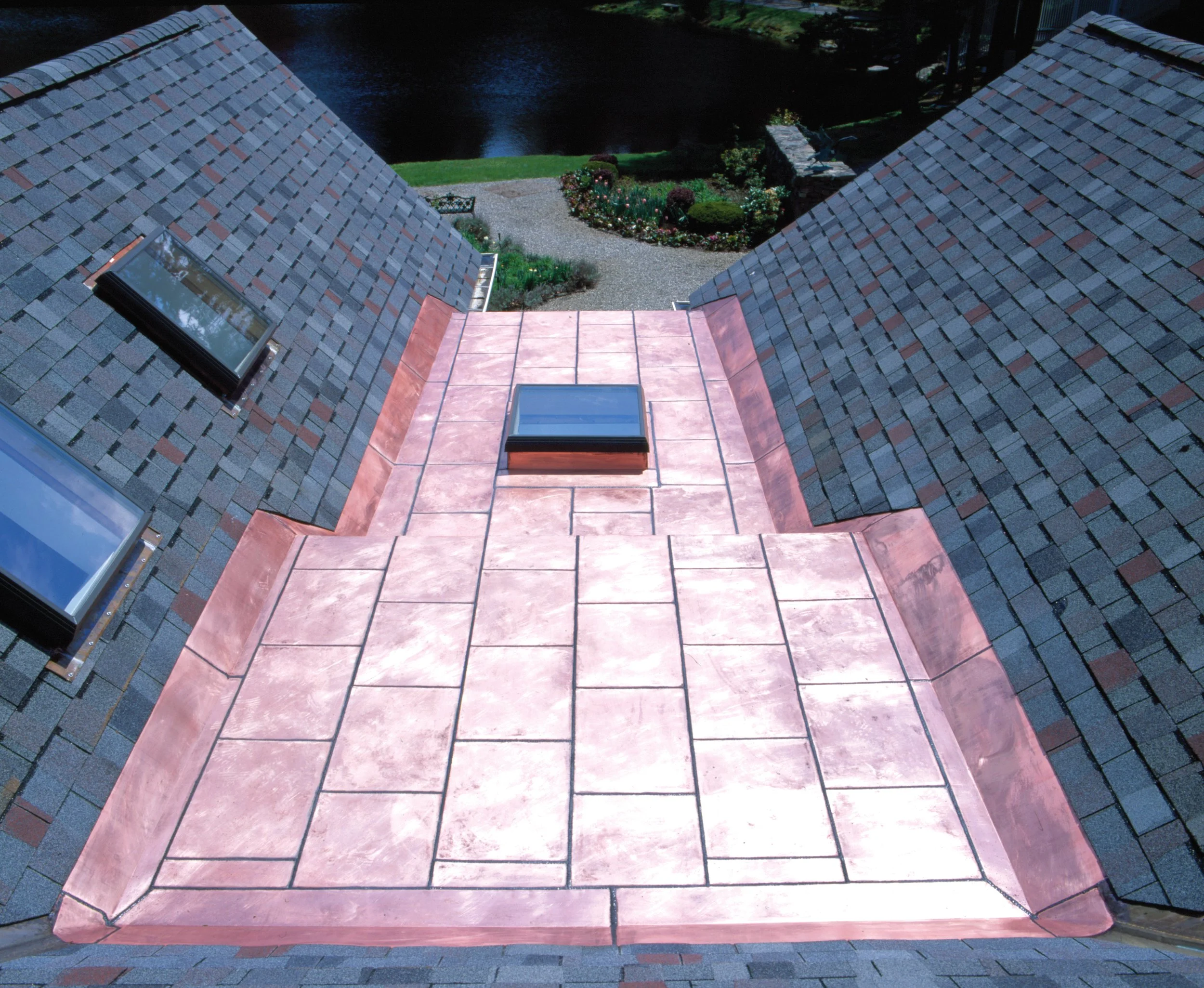 View from a window showing the rooftops of a building with two skylights, leading down to a garden with a pathway, flowers, and bushes, and a body of water in the background.