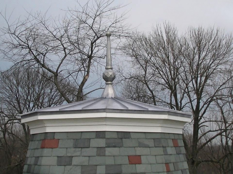 Close-up of a metal-roofed dome with a decorative spire on top, set against bare winter trees and a cloudy sky.