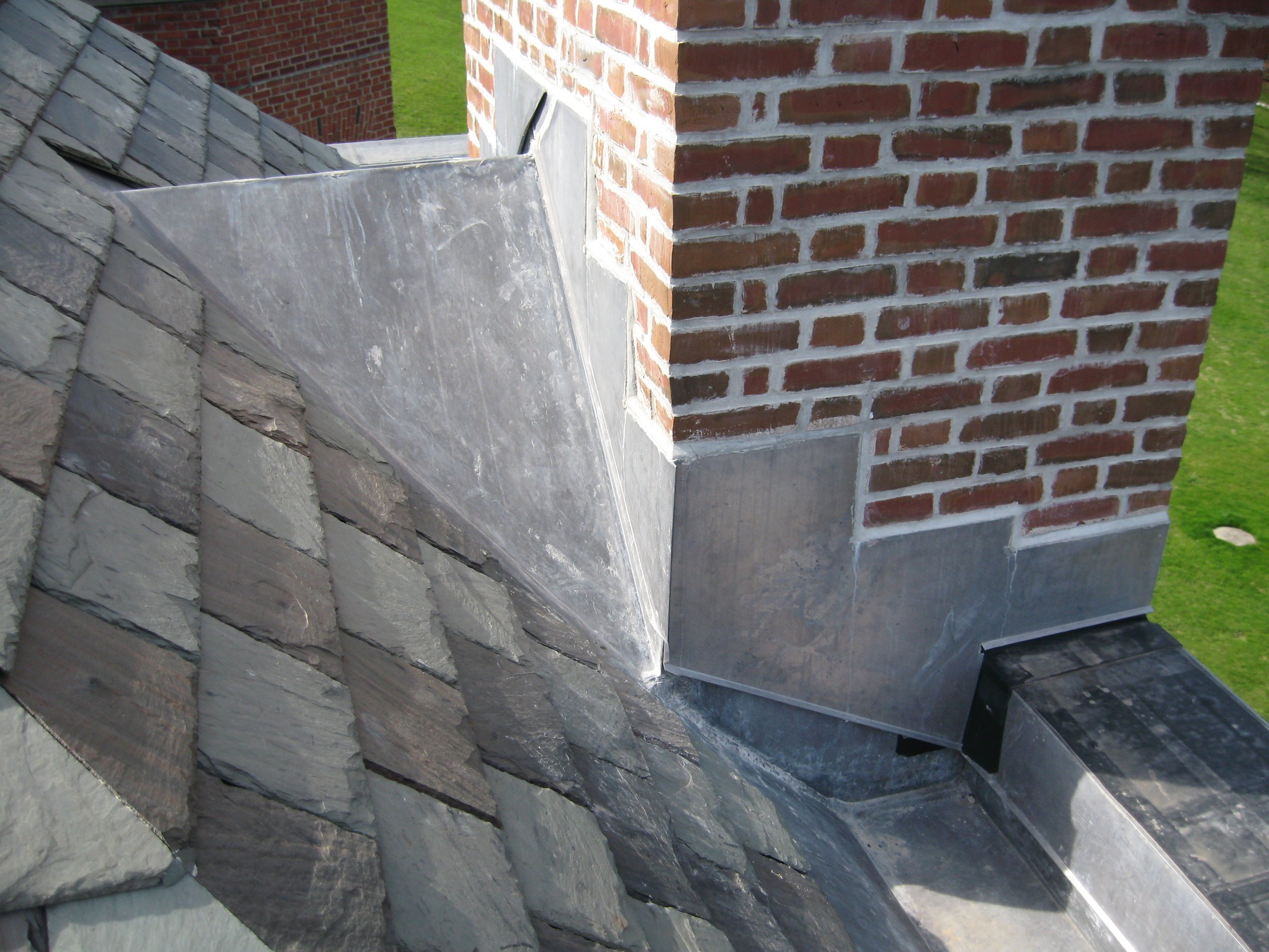 Close-up of a brick chimney with a section of metal flashing and a slate roof, on a building with green grass in the background.